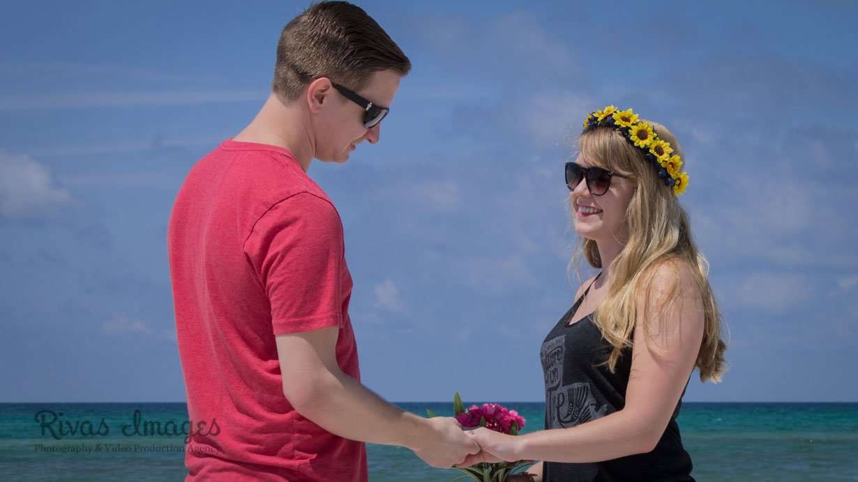 Couple in sunglasses holding hands on the beach, she wears a sunflower crown and holds pink flowers