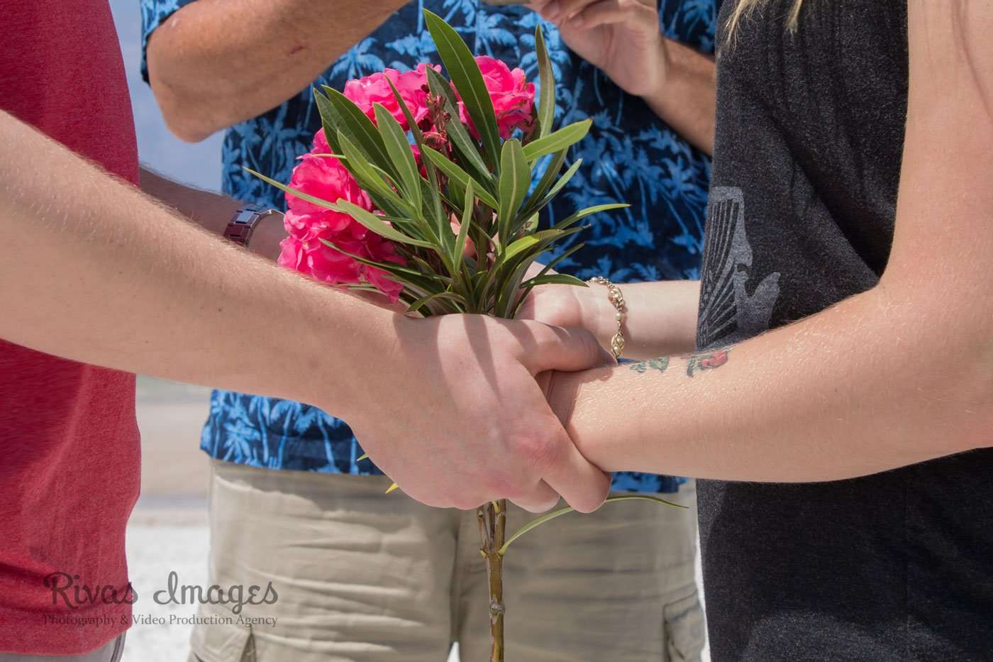 Couple holding hands around a bouquet of pink oleander flowers while an officiant in a blue palm-print shirt stands behind