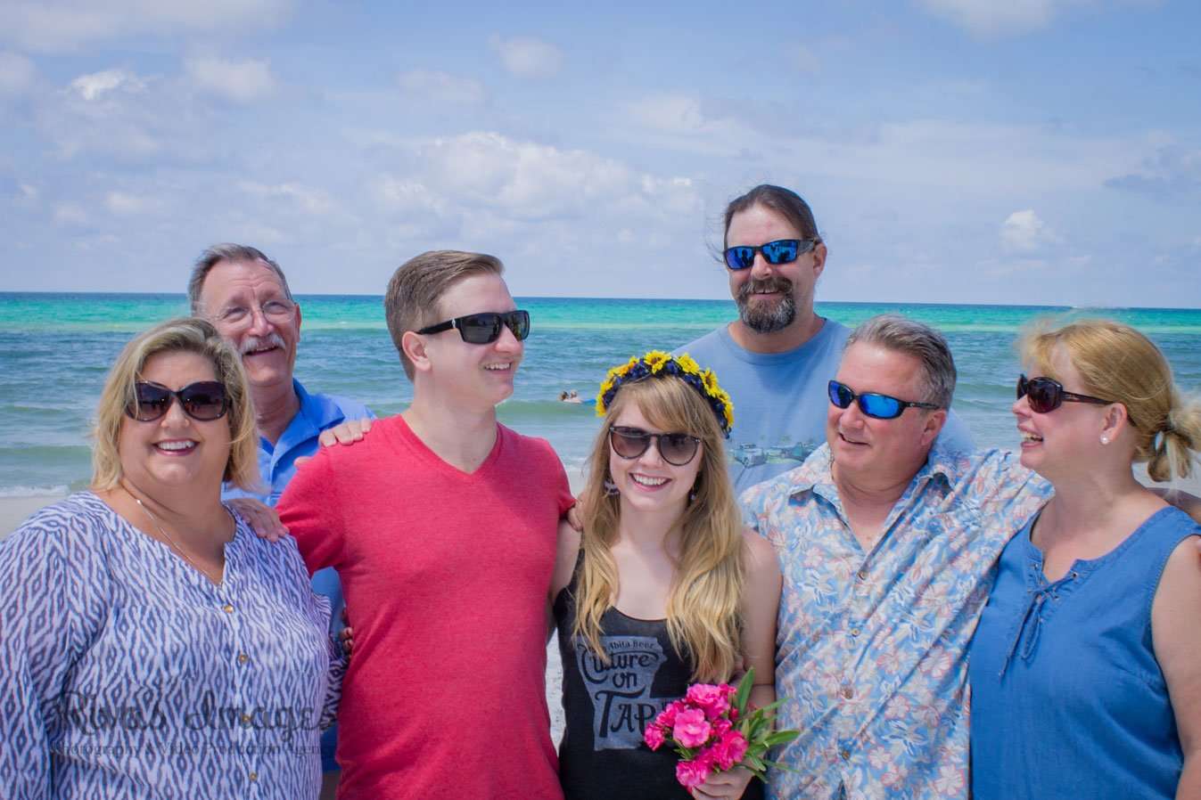 Six smiling people in sunglasses posing close together on the beach, one wearing a flower crown and holding pink flowers