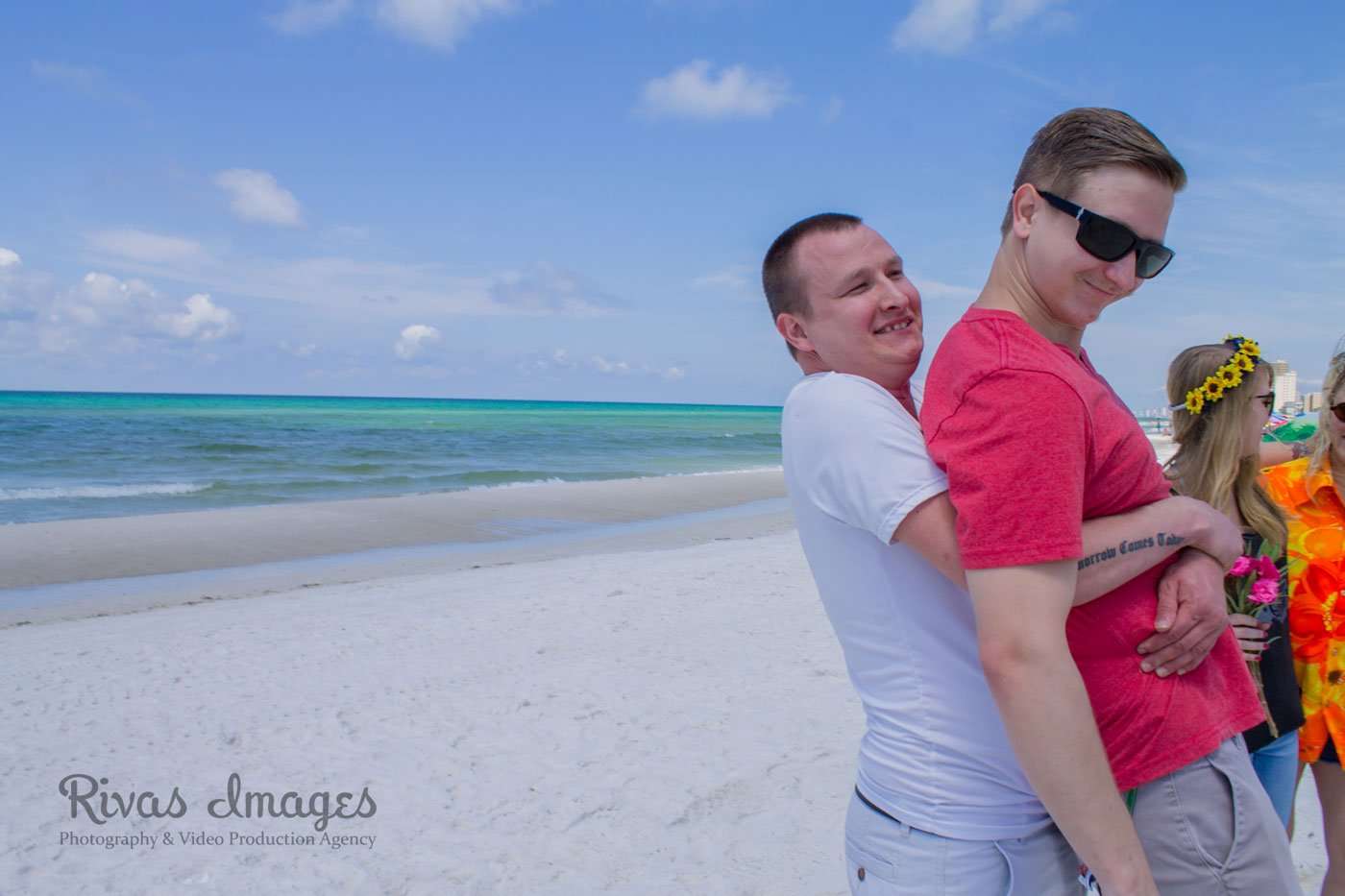 Smiling man hugging a man in a red shirt and sunglasses from behind on a white sand beach by turquoise water