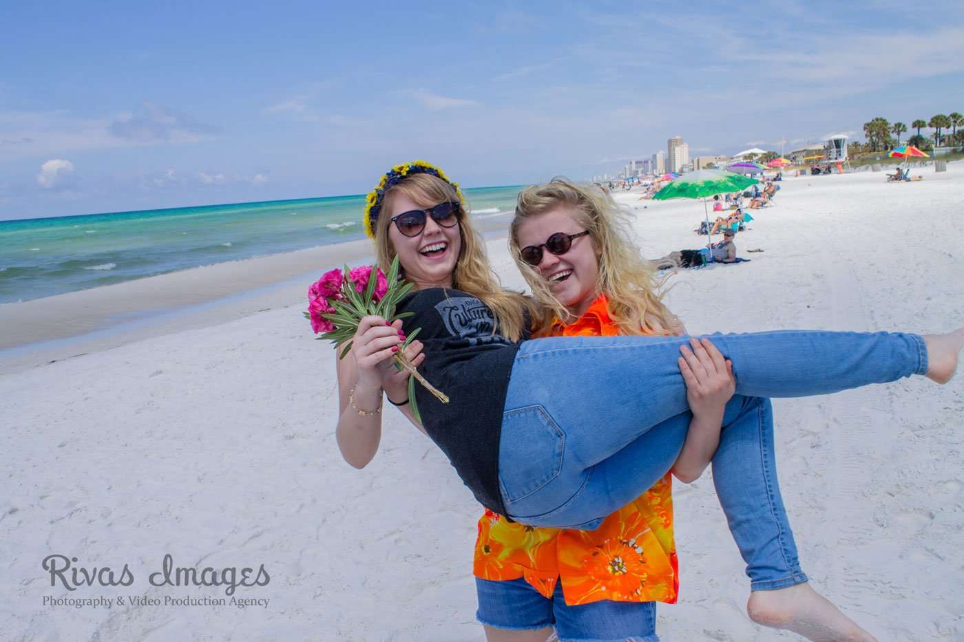 Two laughing women in sunglasses on a white sand beach, one cradling the other while she holds pink flowers