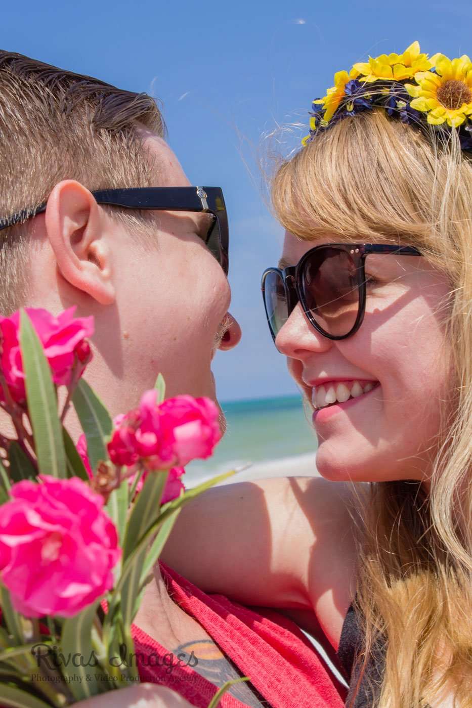 Smiling couple in sunglasses touching noses on the beach, she wears a sunflower crown, he holds pink oleander flowers