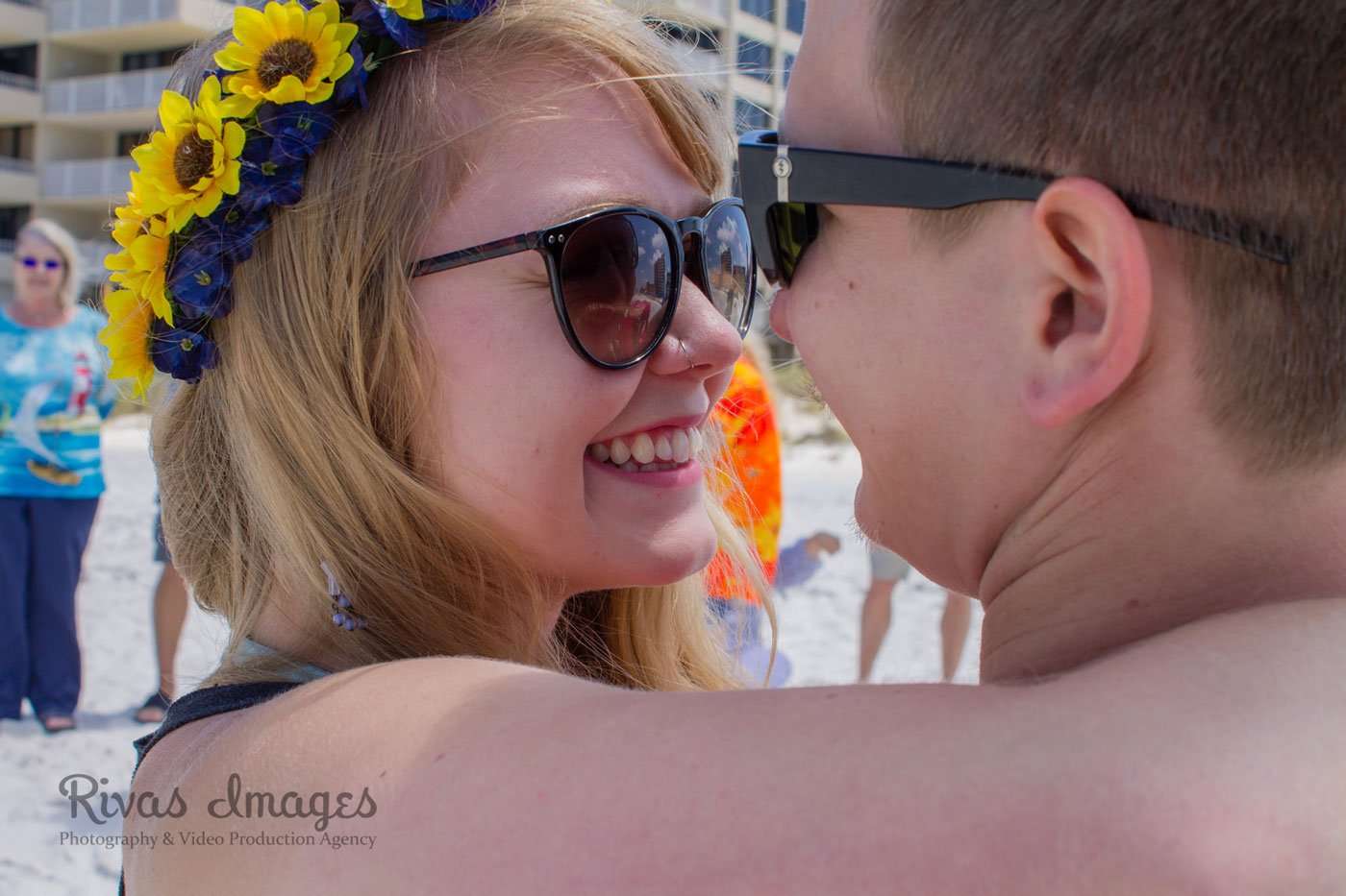 Smiling woman in sunglasses and a sunflower crown embraces her partner on the beach, with people in the background