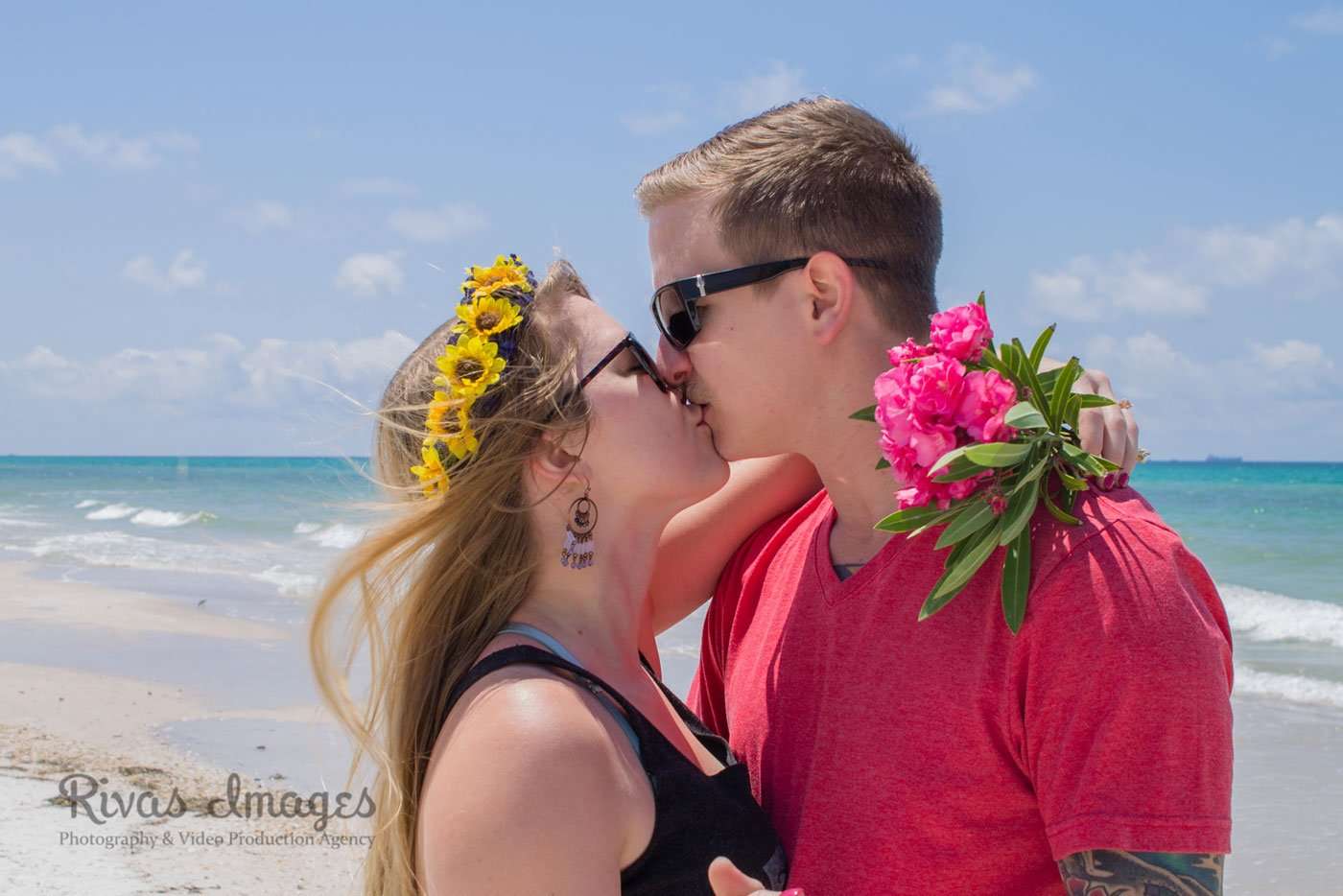 Couple in sunglasses kissing on the beach, she wears a sunflower crown and he holds pink oleander flowers