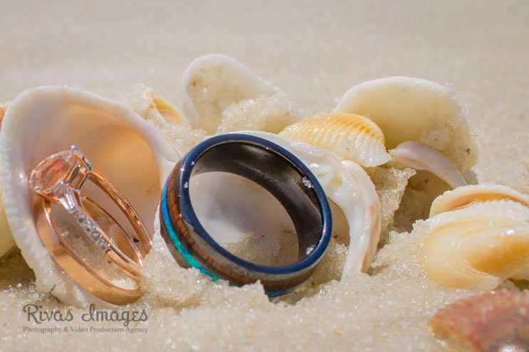 Rose gold diamond engagement ring and a blue-edged wood inlay wedding band nestled among seashells in white sand