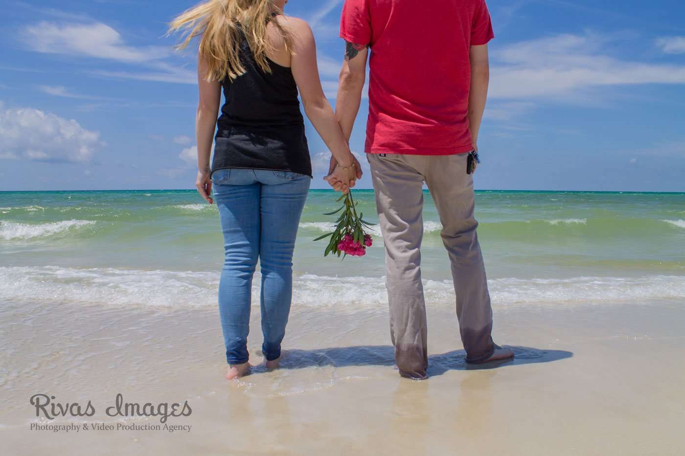 Couple seen from behind holding hands and a pink flower bouquet while standing barefoot on the beach by turquoise water