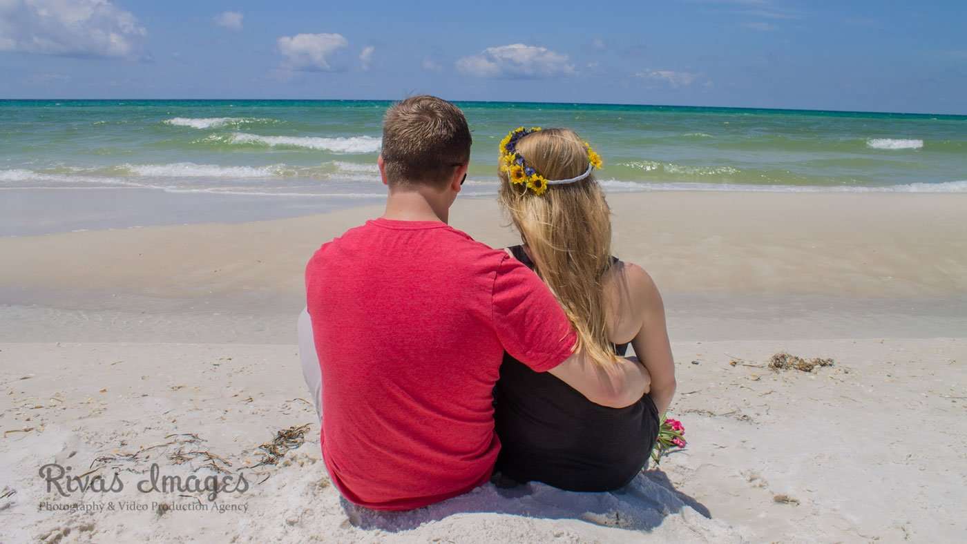 Couple sitting close on white sand facing the turquoise water, her hair wearing a yellow flower crown