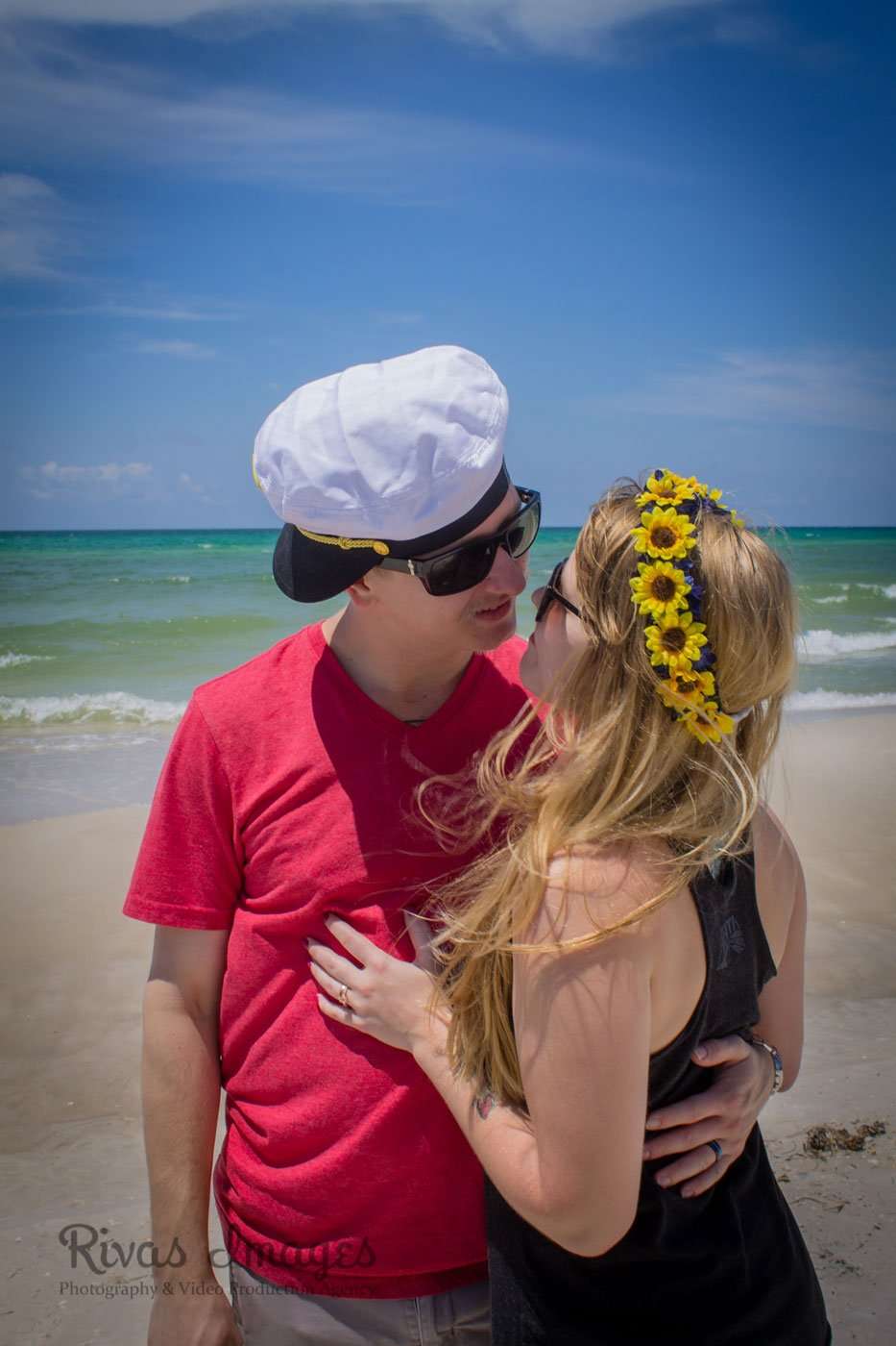 Couple embracing on the beach, he in a white captain's hat, she wearing a sunflower crown, turquoise water behind