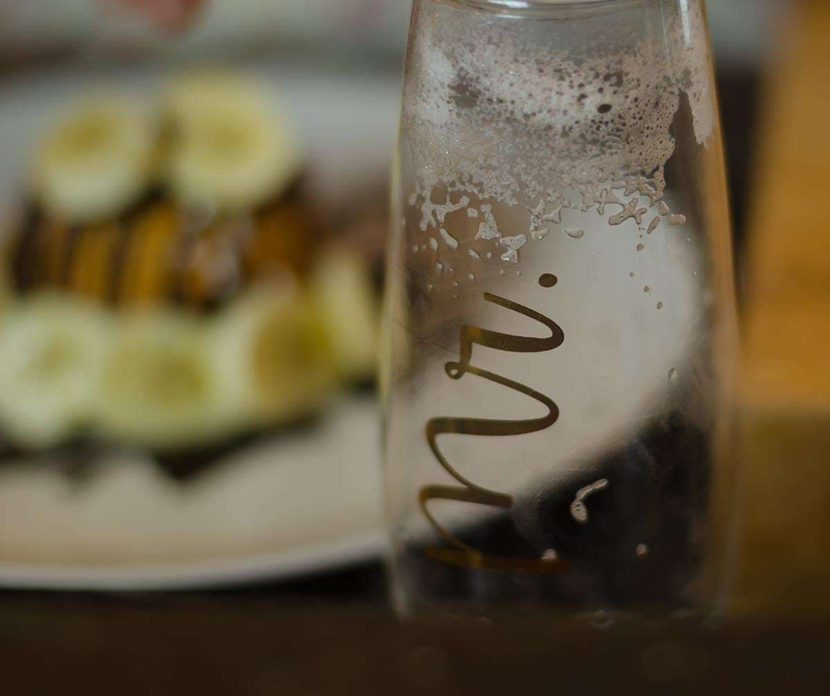 Empty glass bottle with gold "Mr." script lettering beside a blurred plate of banana slices and chocolate drizzle