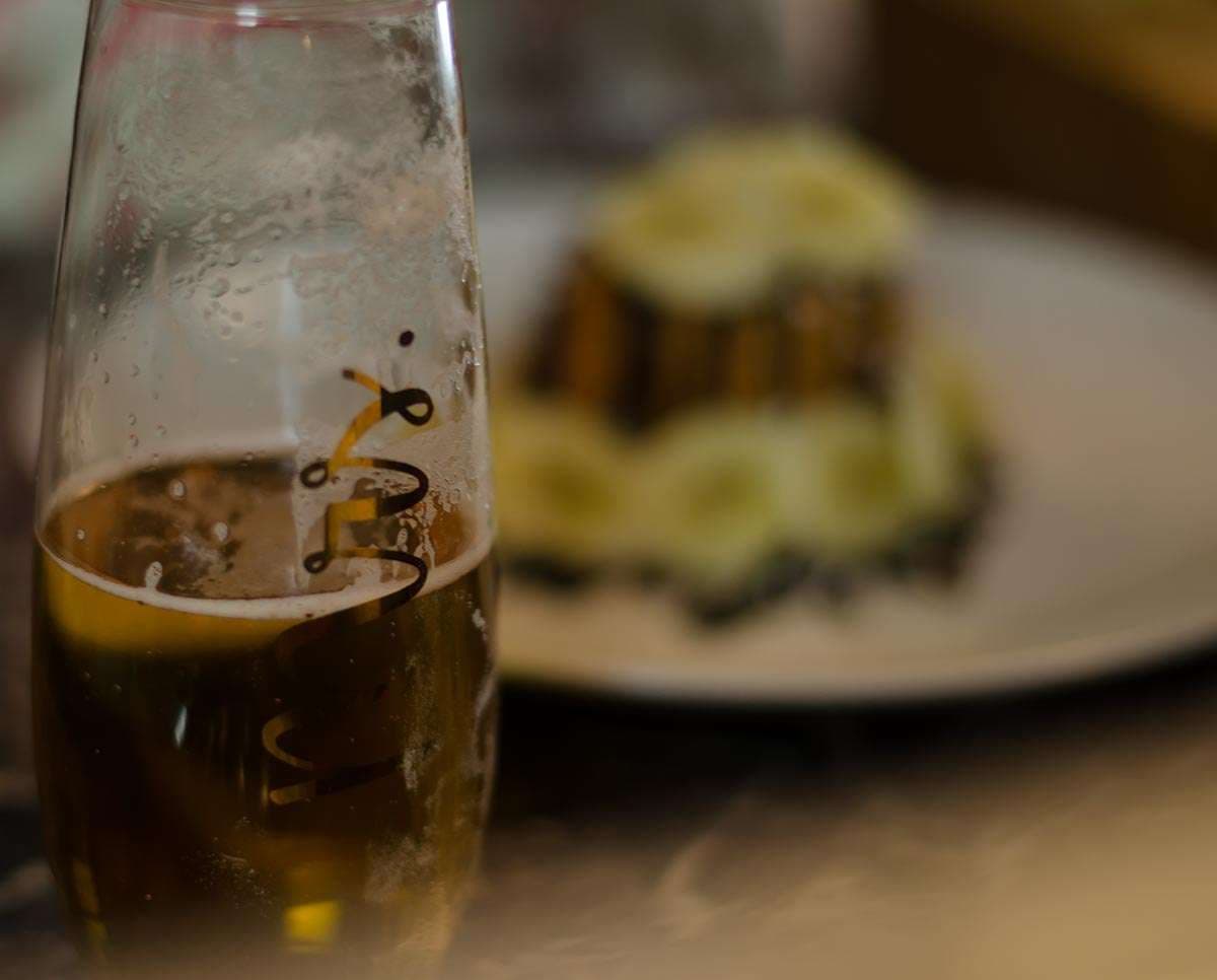 Close-up of a condensation-covered glass of golden beer beside a blurred plated dessert on a white plate