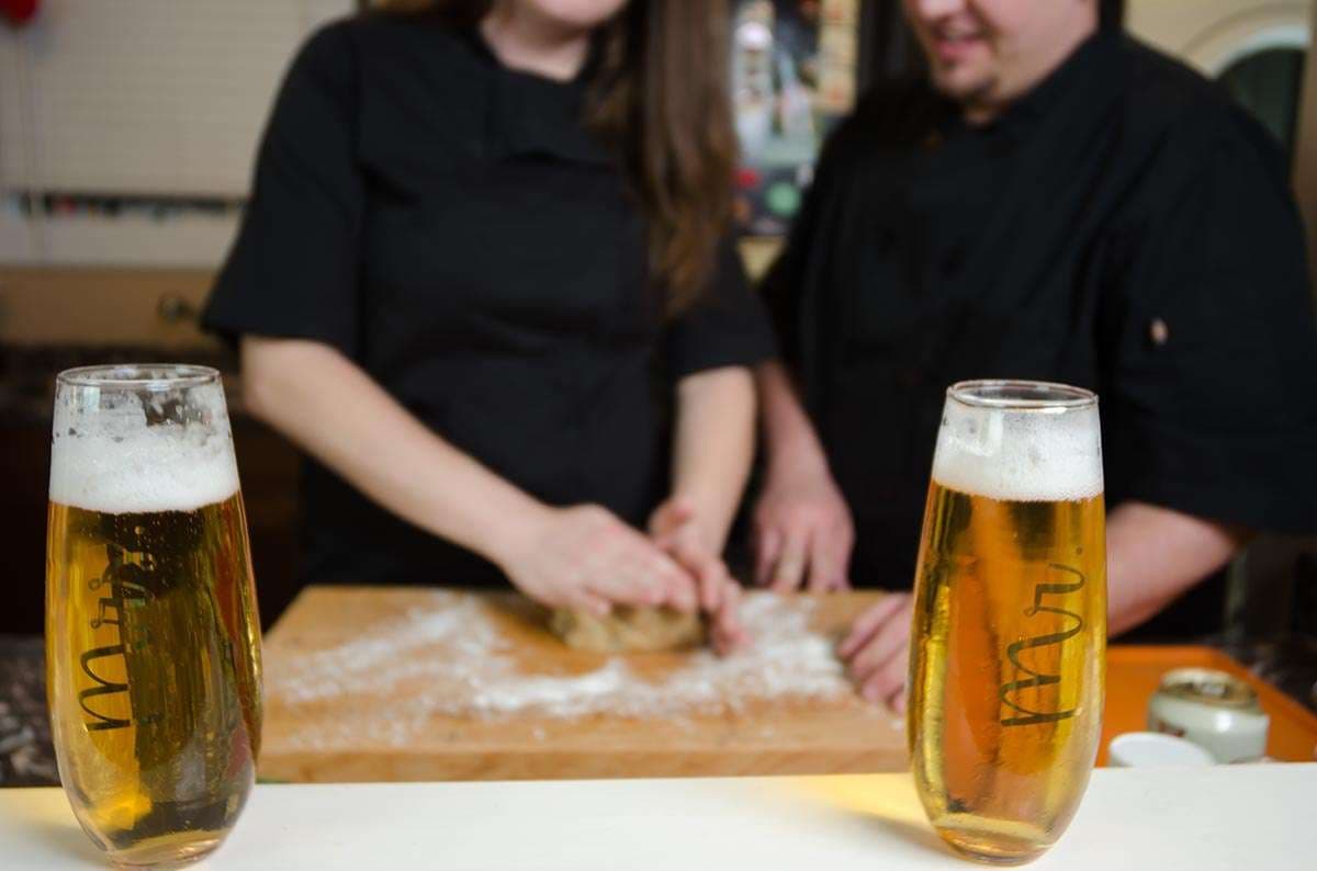 Couple in black chef coats kneading dough on a floured wooden board behind two glasses of beer