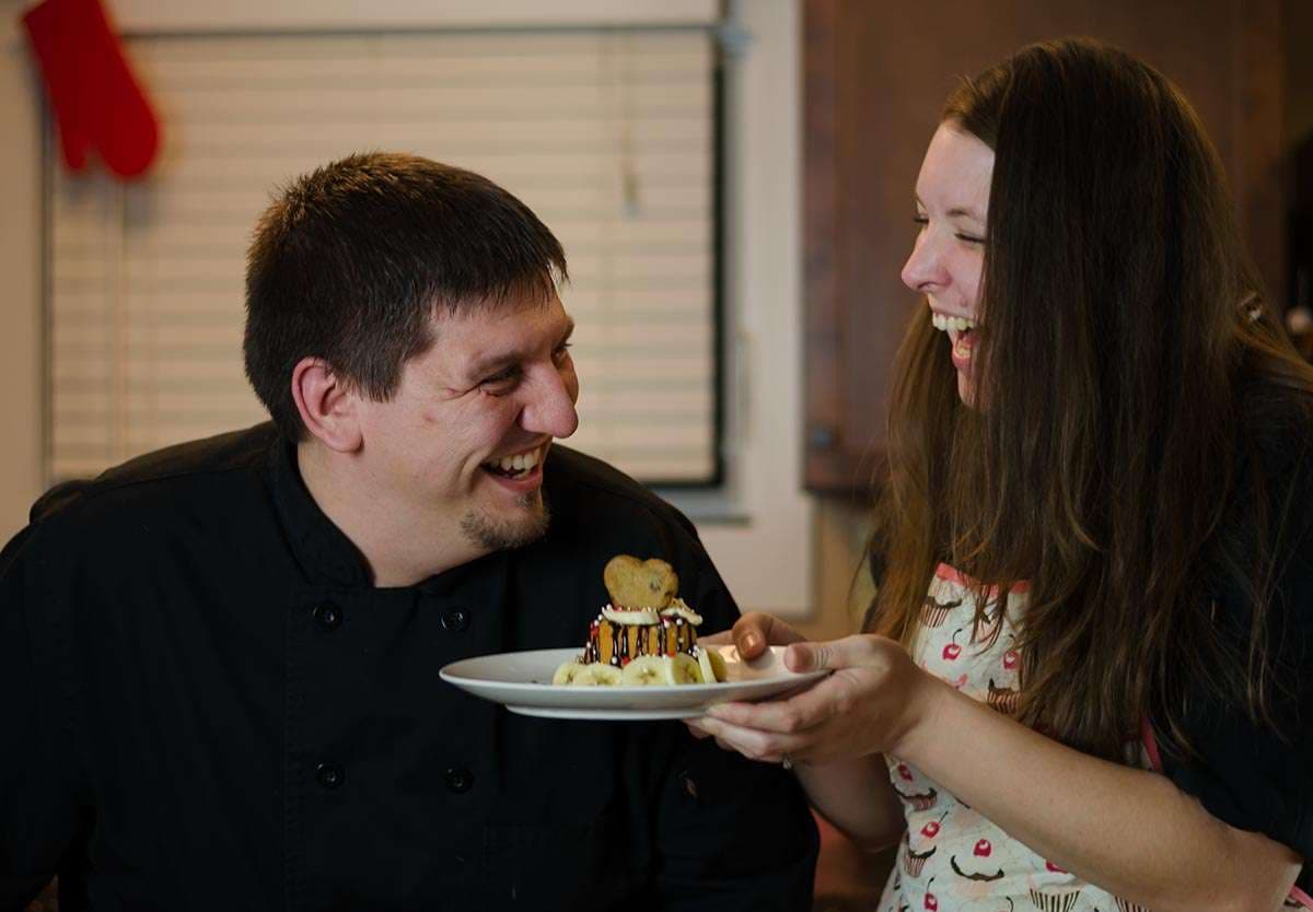 Laughing chef in black jacket and woman in cupcake apron share a small decorated dessert on a plate in a kitchen