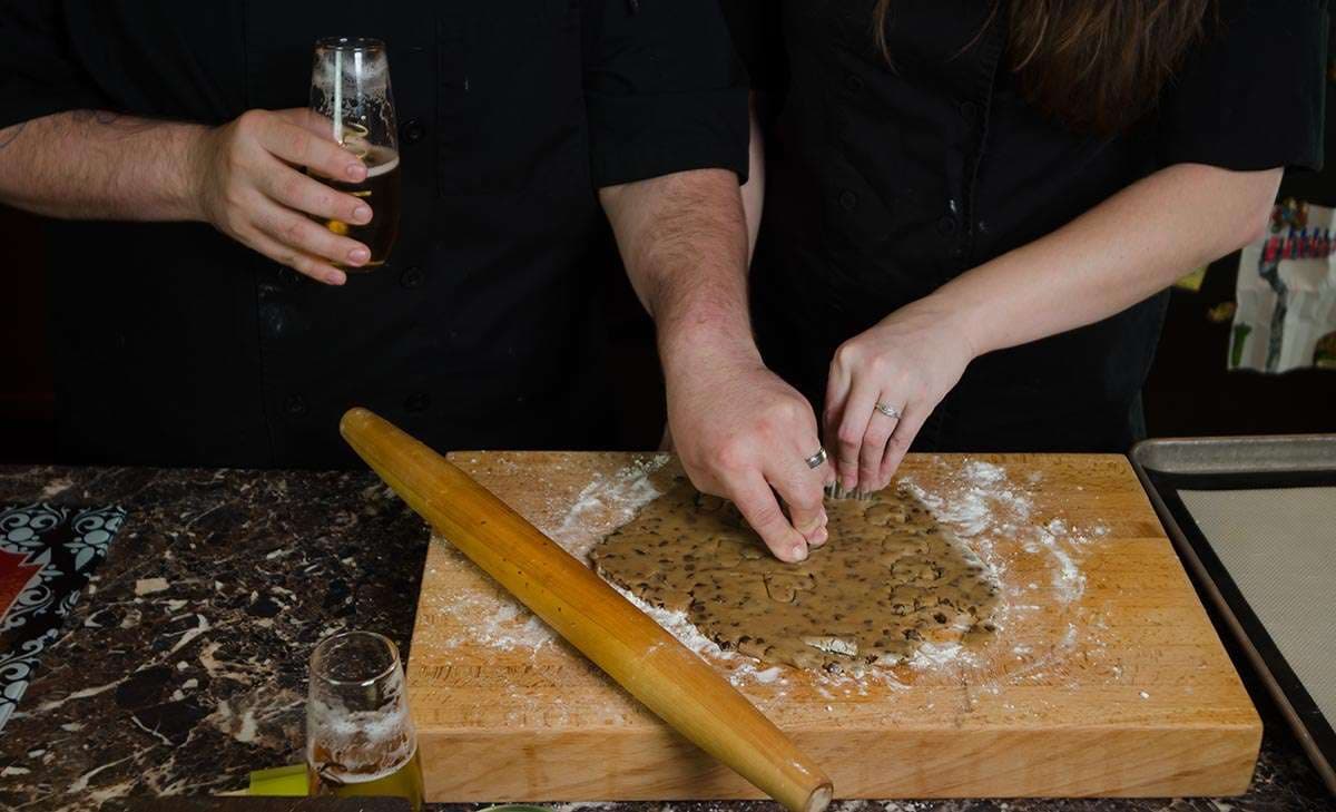 Couple pressing cookie dough on a floured wooden board beside a rolling pin and glasses of beer
