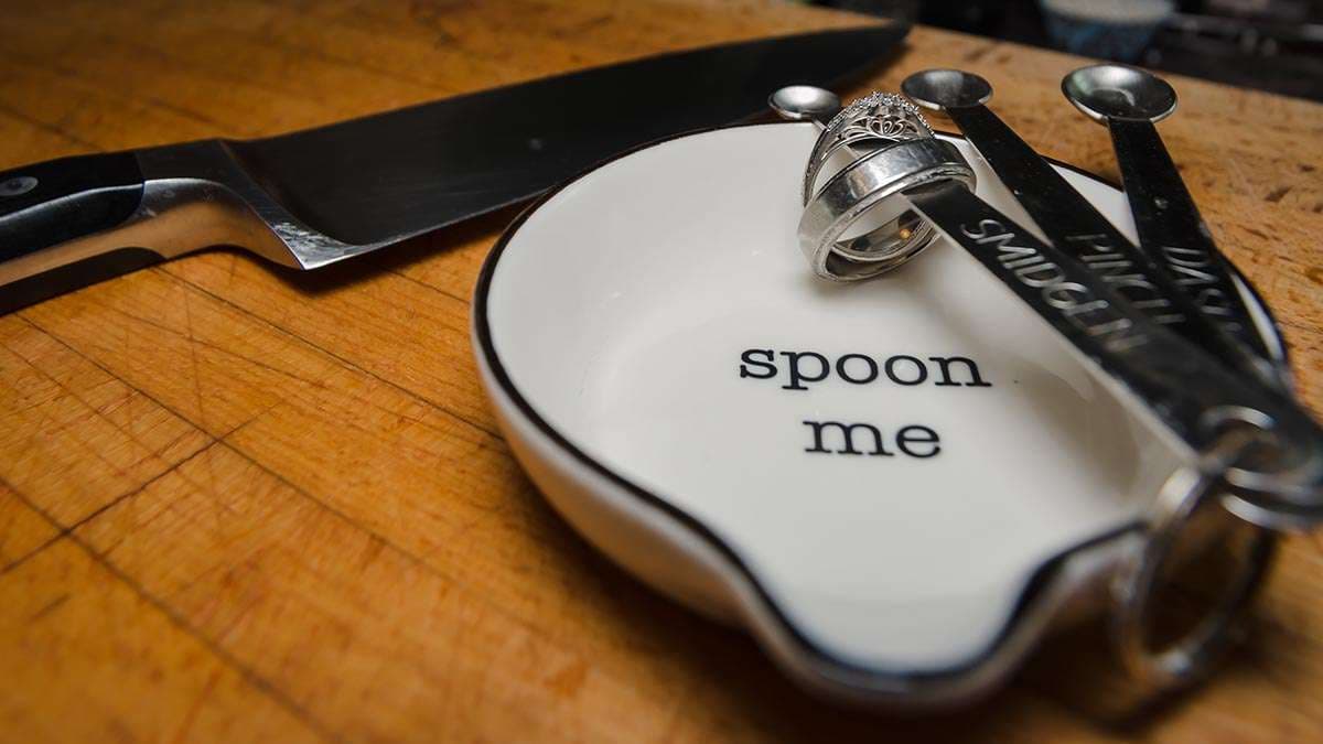 Silver ring resting on a white spoon rest reading "spoon me" beside measuring spoons and a chef's knife on a wooden board