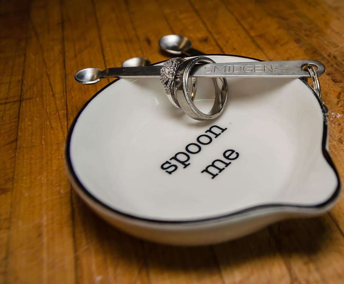 Two silver rings resting on a Smidgen measuring spoon over a white dish printed with "spoon me" on a wooden counter