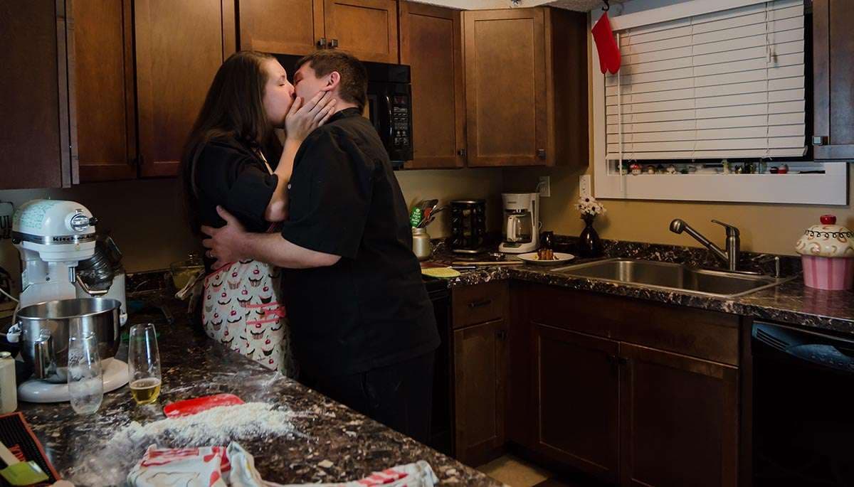 Couple kissing in a home kitchen beside a stand mixer and a flour-dusted granite counter with a beer glass