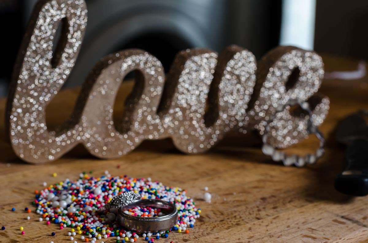 Two rings resting in a pile of colorful sprinkles on a wooden board in front of a glittery gold "love" sign