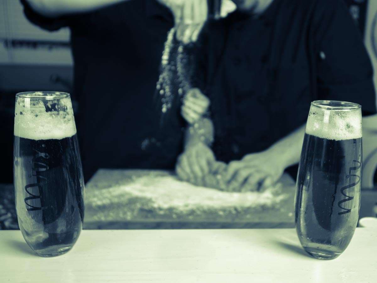 Couple kneading dough on a floured board behind two glasses of dark beer, in a blue-green tinted image