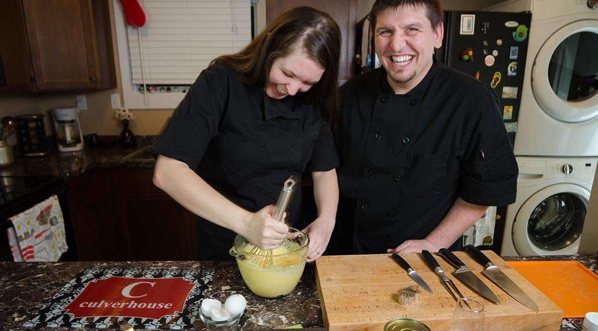 Couple in black chef coats laughing in a kitchen as she whisks a yellow mixture beside knives on a cutting board
