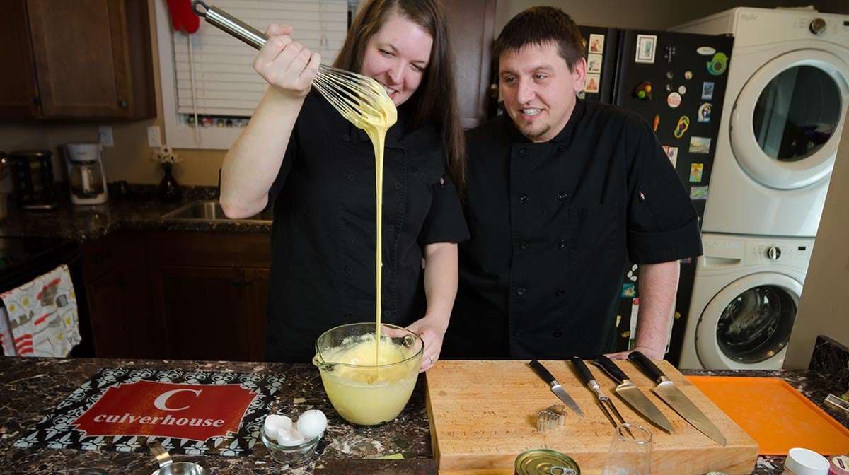 Couple in black chef coats in a home kitchen, one whisking yellow sauce in a glass bowl while the other watches