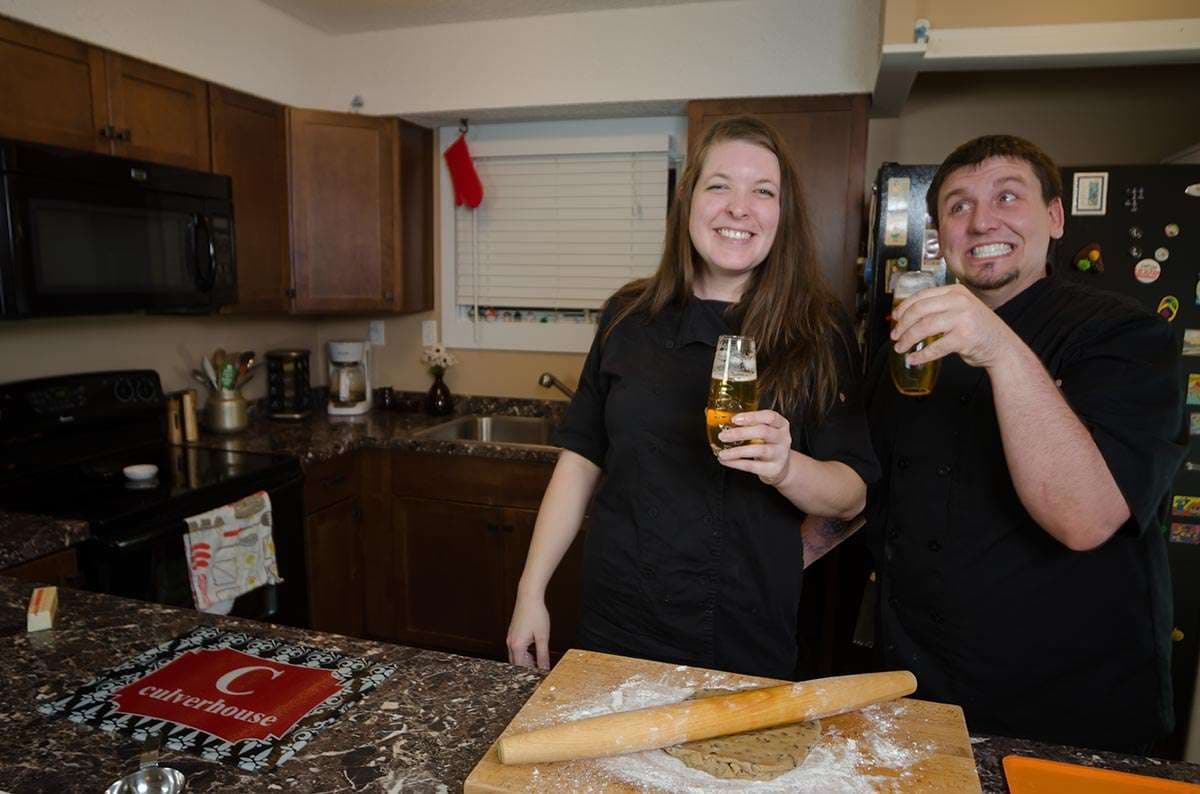 Smiling couple in black chef jackets hold beer glasses in a home kitchen beside a floured board and rolling pin