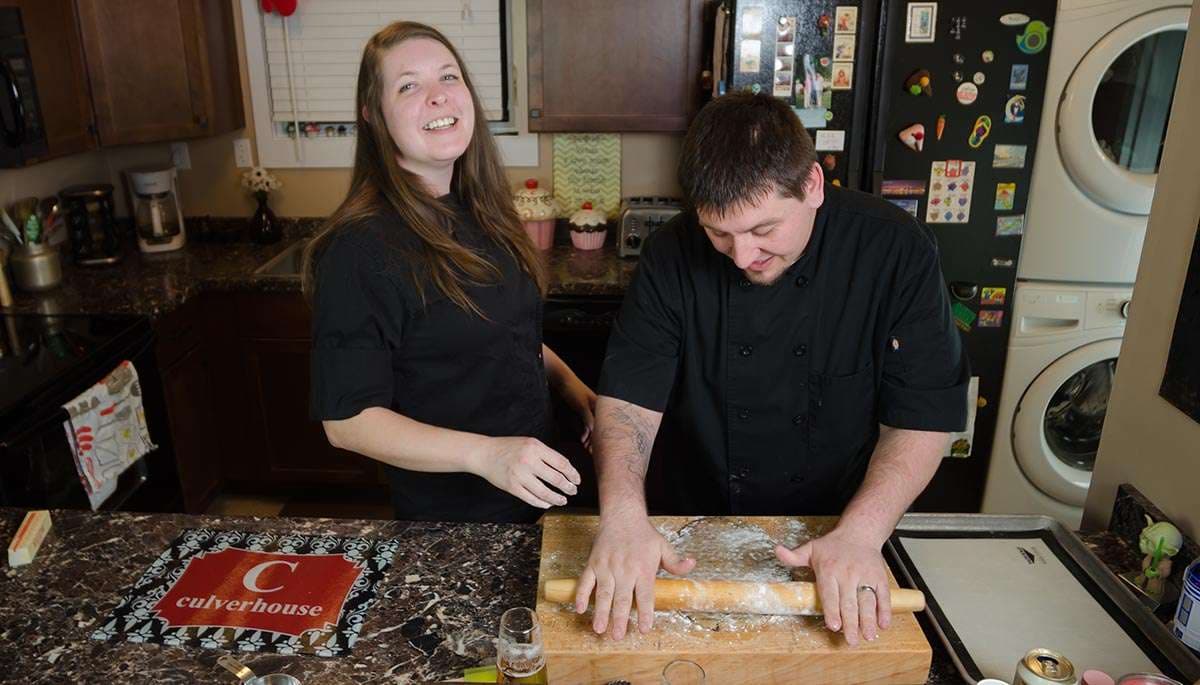 Couple in black chef jackets in a kitchen, she laughing while he rolls dough on a floured wooden board