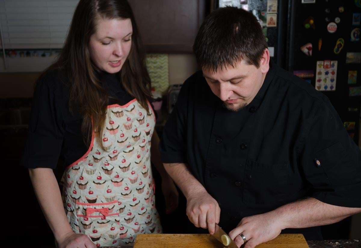 Couple in a kitchen, man in black chef jacket slicing on a wooden board while woman in cupcake apron watches