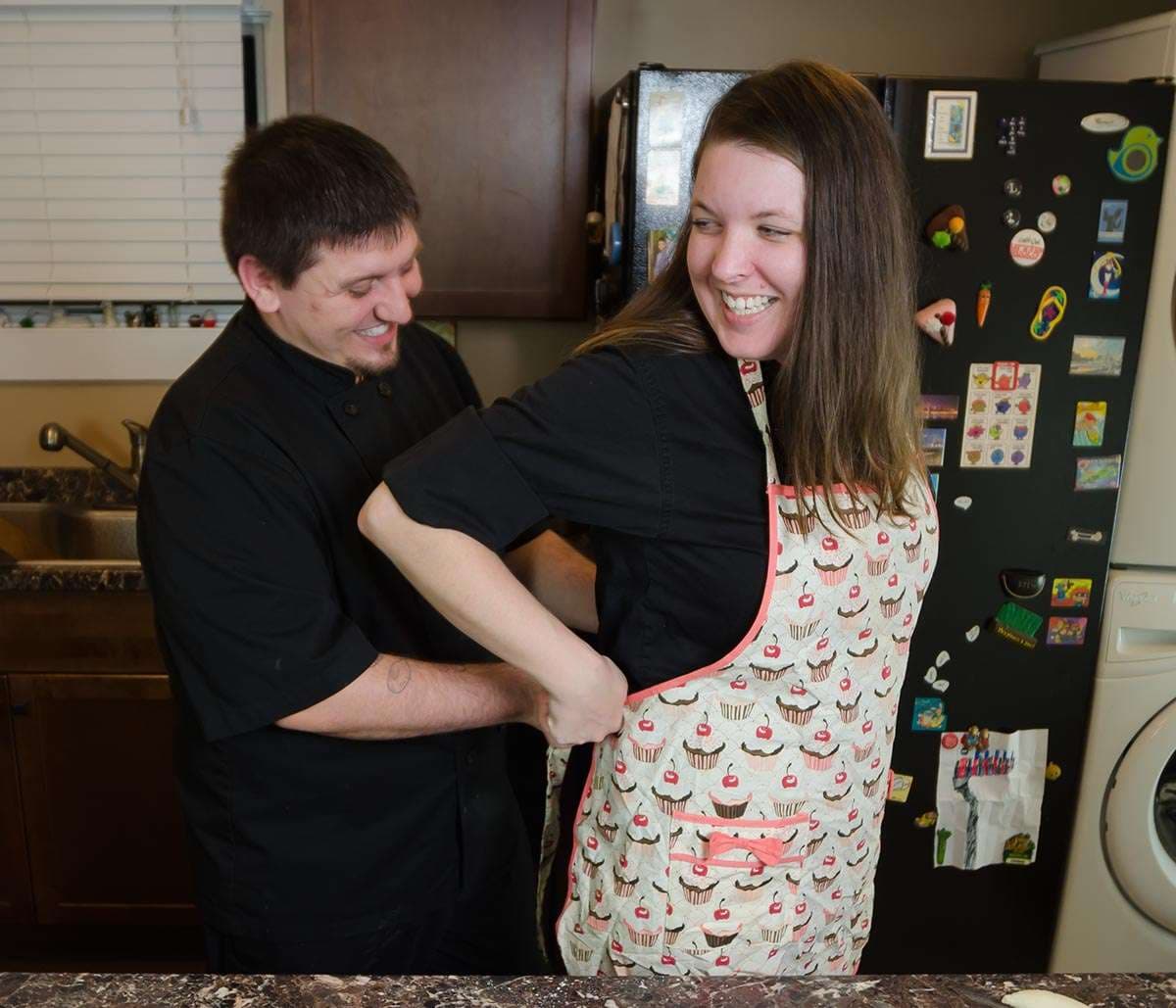 Smiling man in black chef jacket helps a laughing woman tie a cupcake-print apron in a kitchen