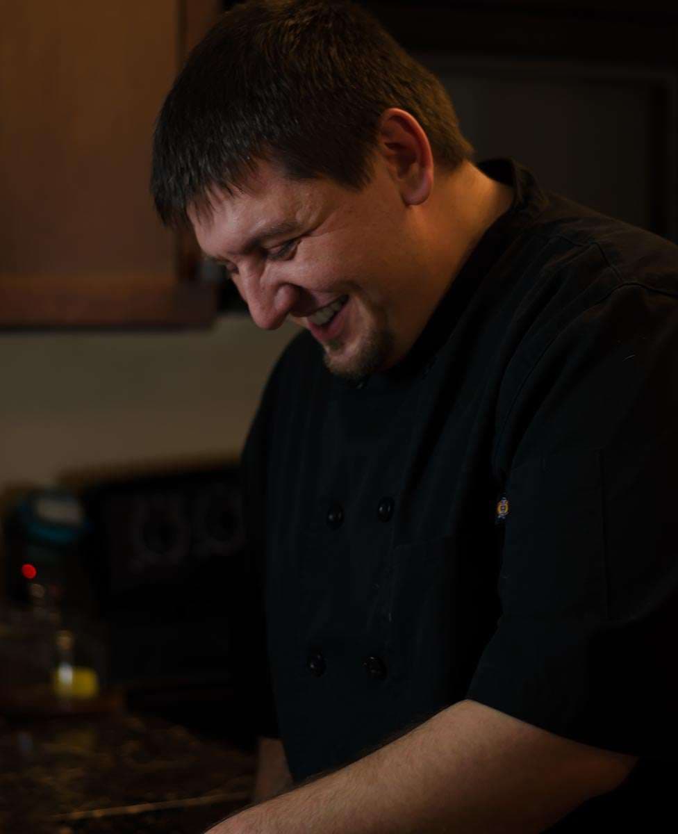Smiling chef in a black double-breasted coat looking down while working at a kitchen counter in warm, low light
