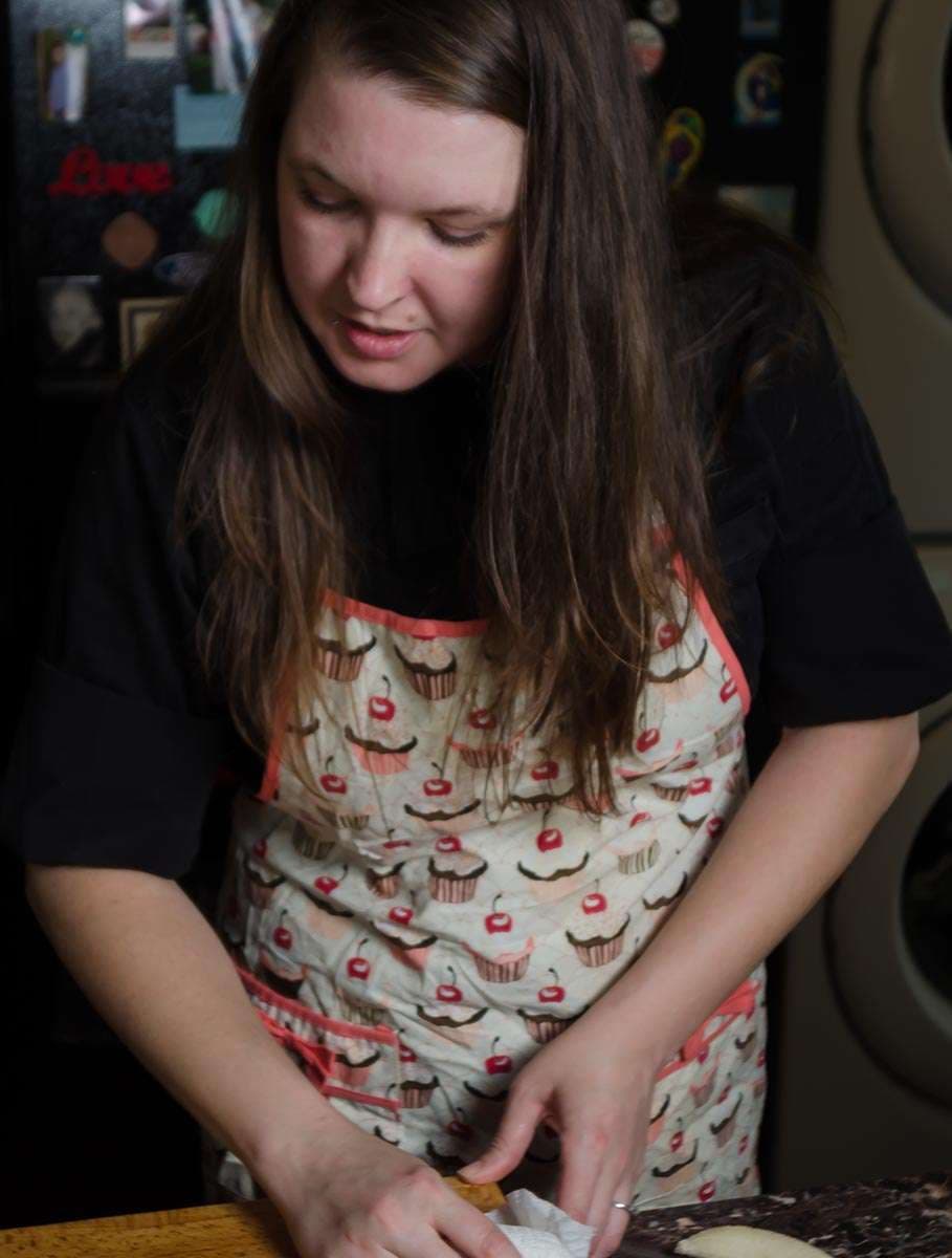 Woman in a cupcake-print apron looking down while working dough on a wooden cutting board in a kitchen