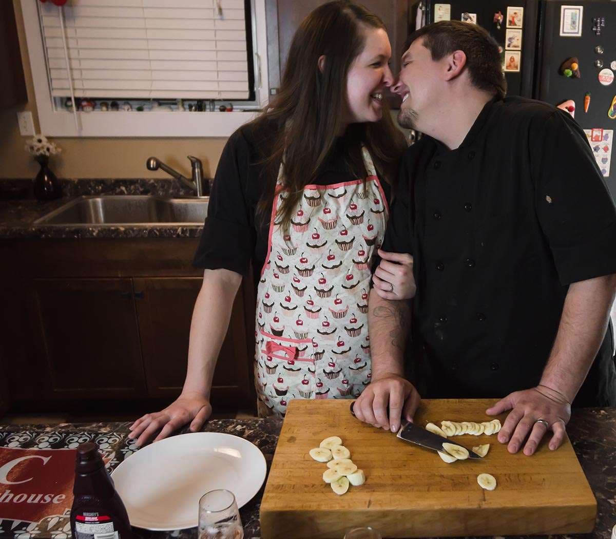 Couple laughing nose to nose in a kitchen while slicing bananas on a wooden board, she wears a cupcake apron