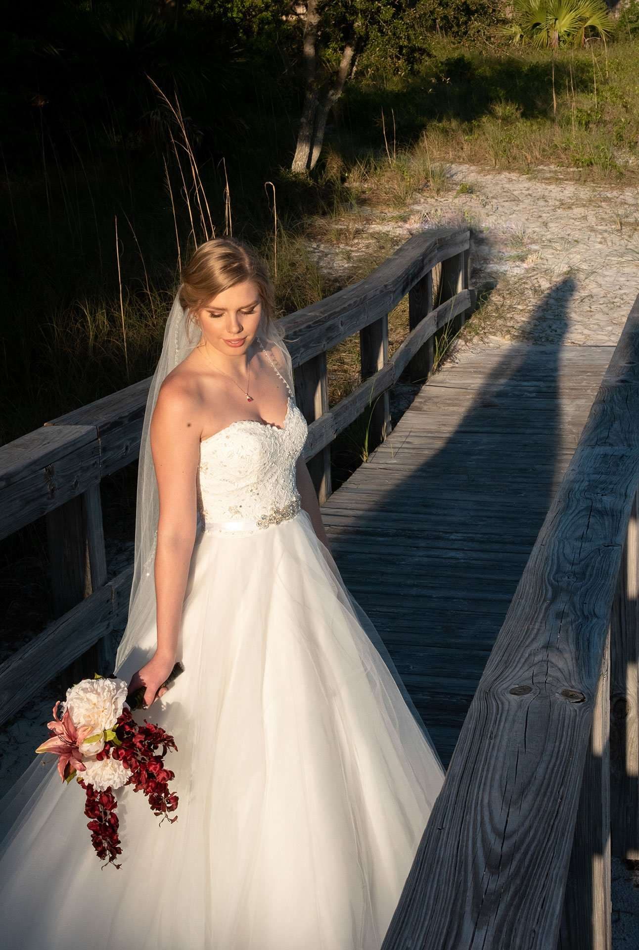 Bride in a strapless white gown and veil holding a red and white bouquet on a wooden boardwalk in evening light