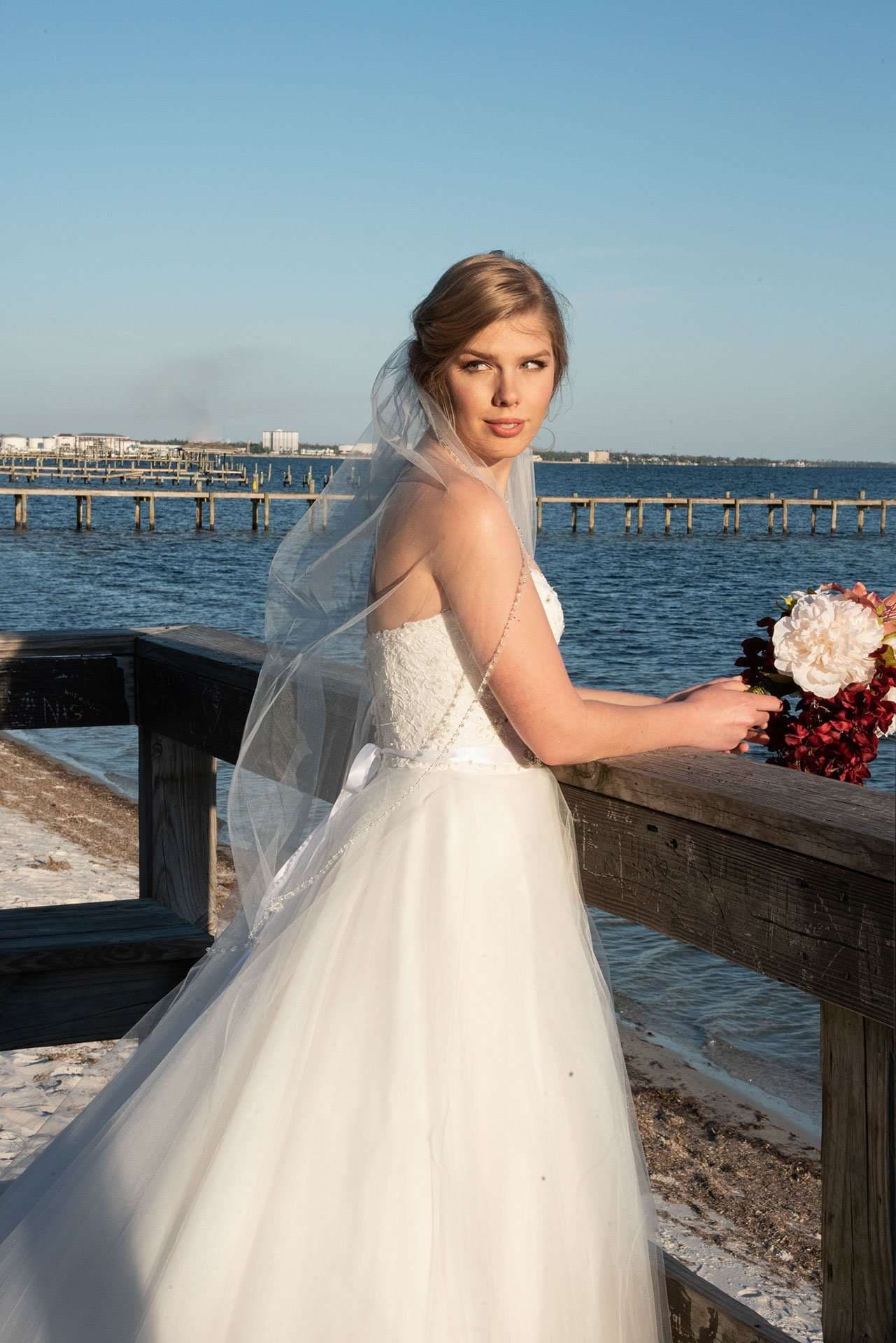 Bride in a strapless white gown and long veil holding a red and white bouquet on a wooden railing by the bay