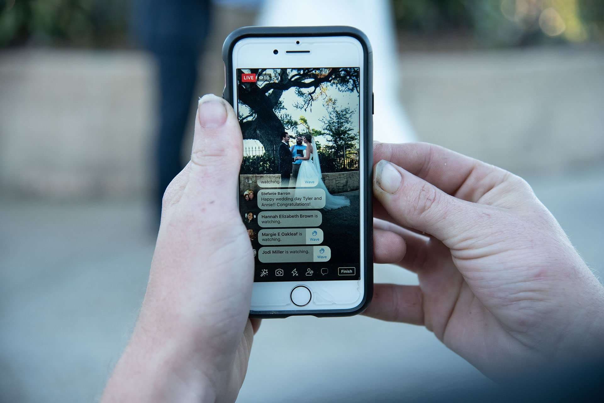 Hands holding a phone that livestreams an outdoor wedding ceremony, with viewer comments shown on screen