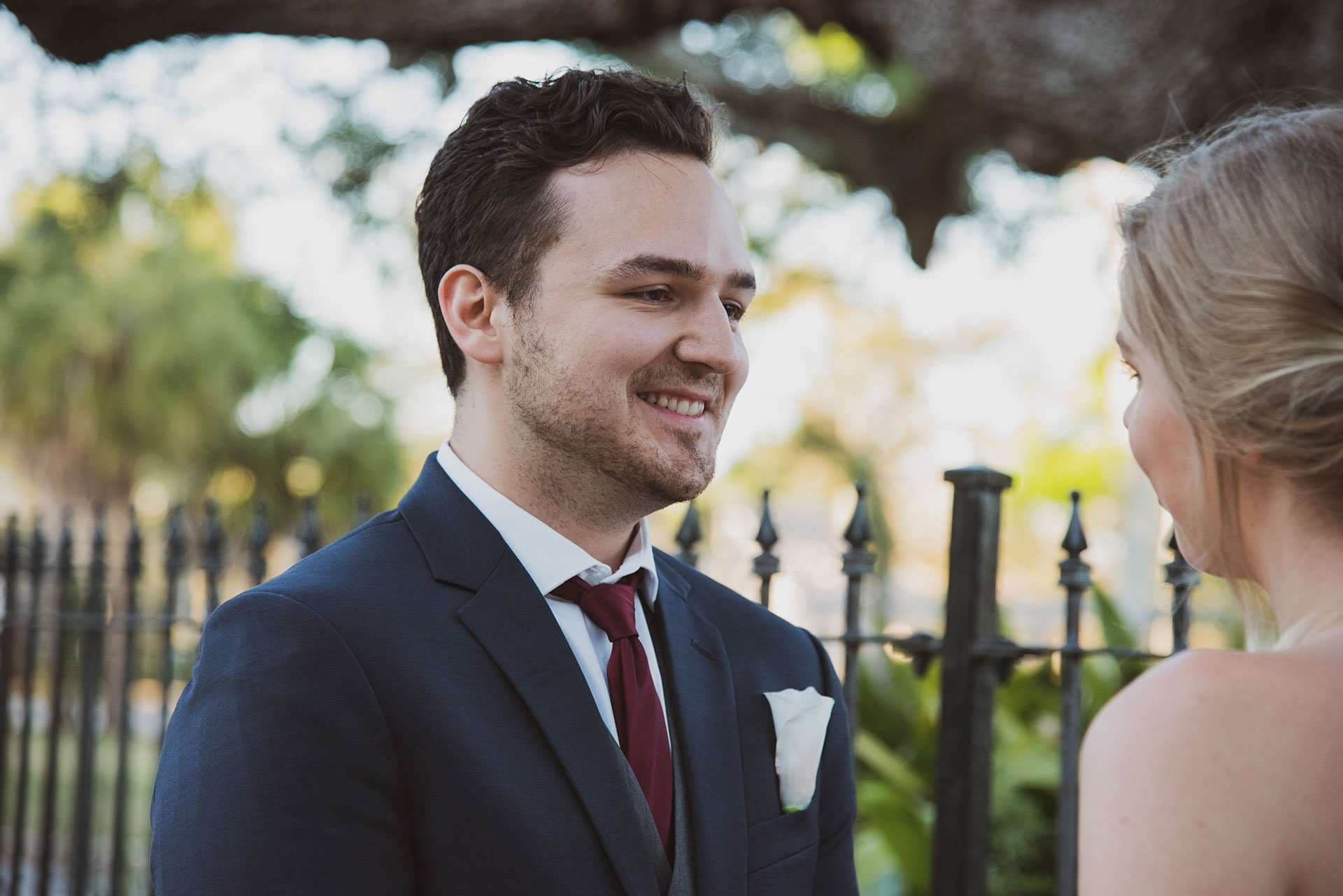 Smiling groom in a navy suit and burgundy tie looks at his bride beside an iron fence under oak branches
