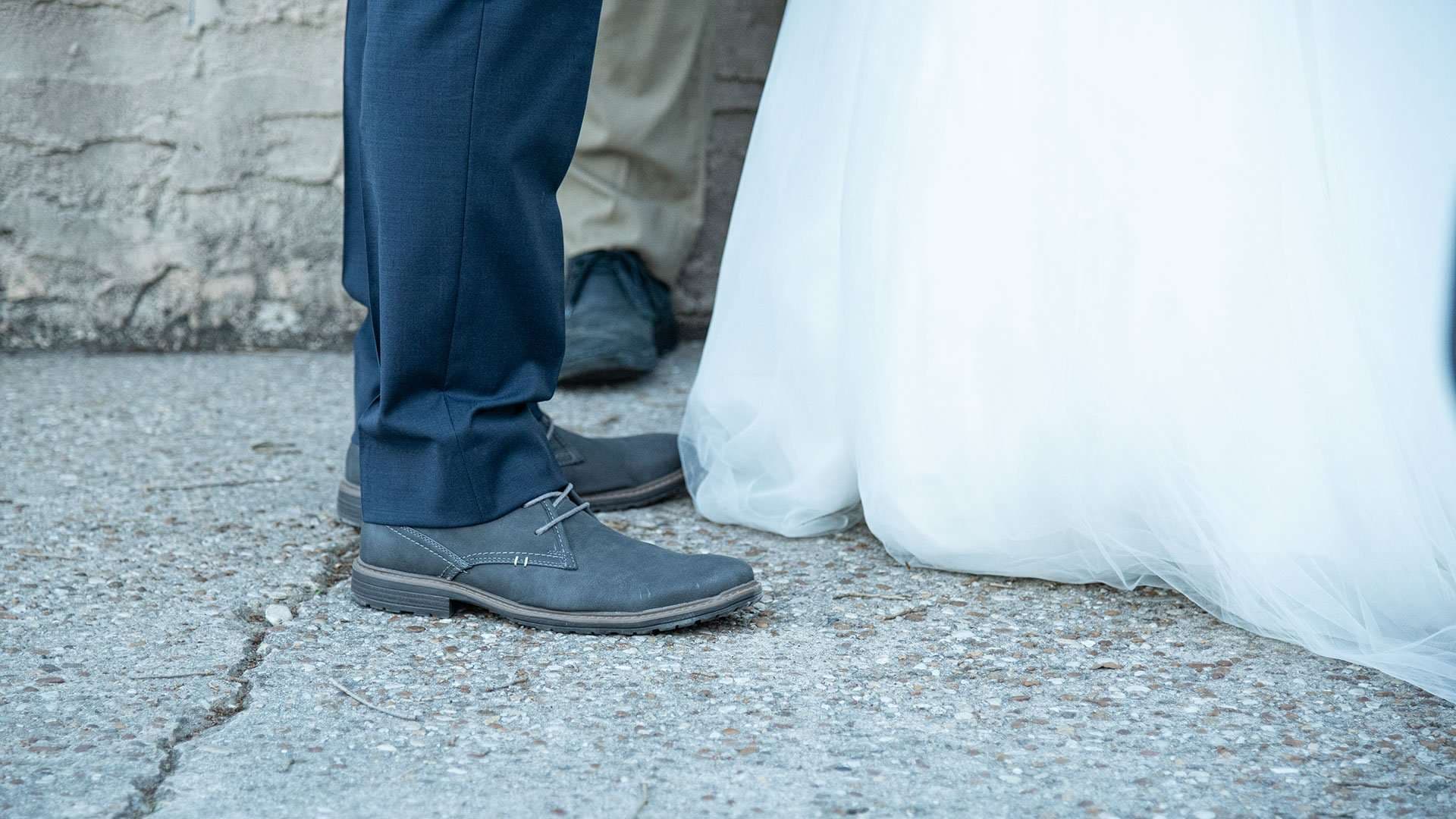 Close-up of a groom's gray suede shoes and navy trousers beside the tulle hem of a bride's white gown