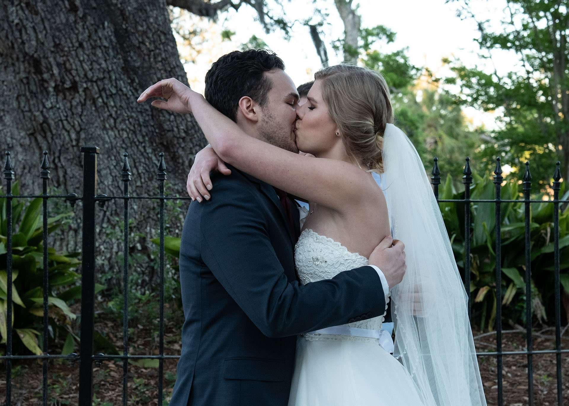 Bride in a lace gown and veil kissing her groom in a navy suit beside an iron fence and large oak tree