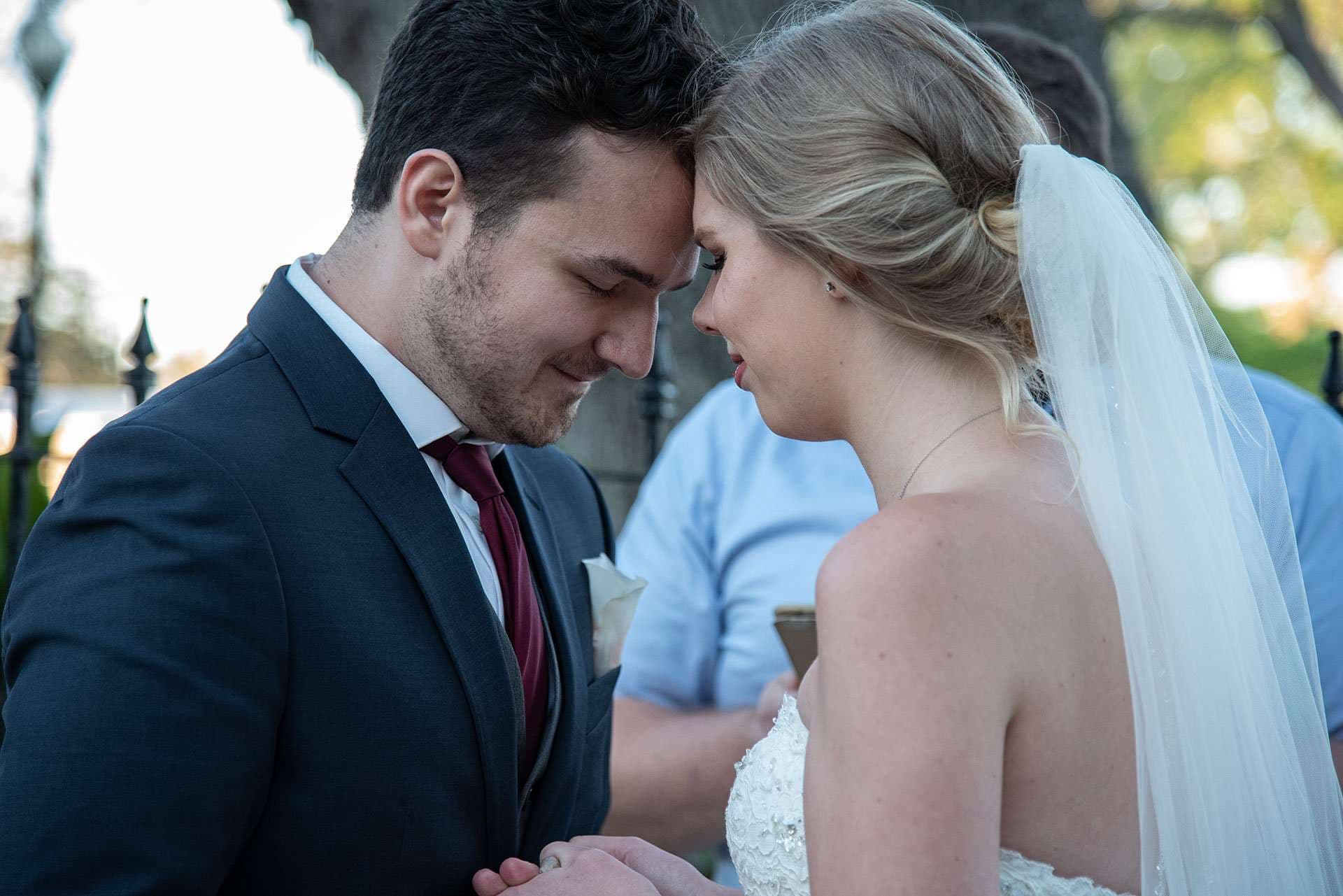 Couple touching foreheads with eyes closed, holding hands outdoors, bride in a lace gown and long veil, groom in navy suit