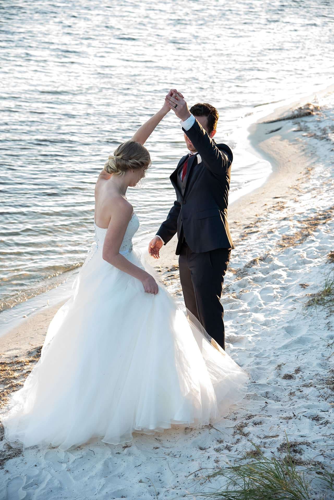 Couple dancing on a sandy shoreline as he twirls his bride in a white tulle gown beside the water