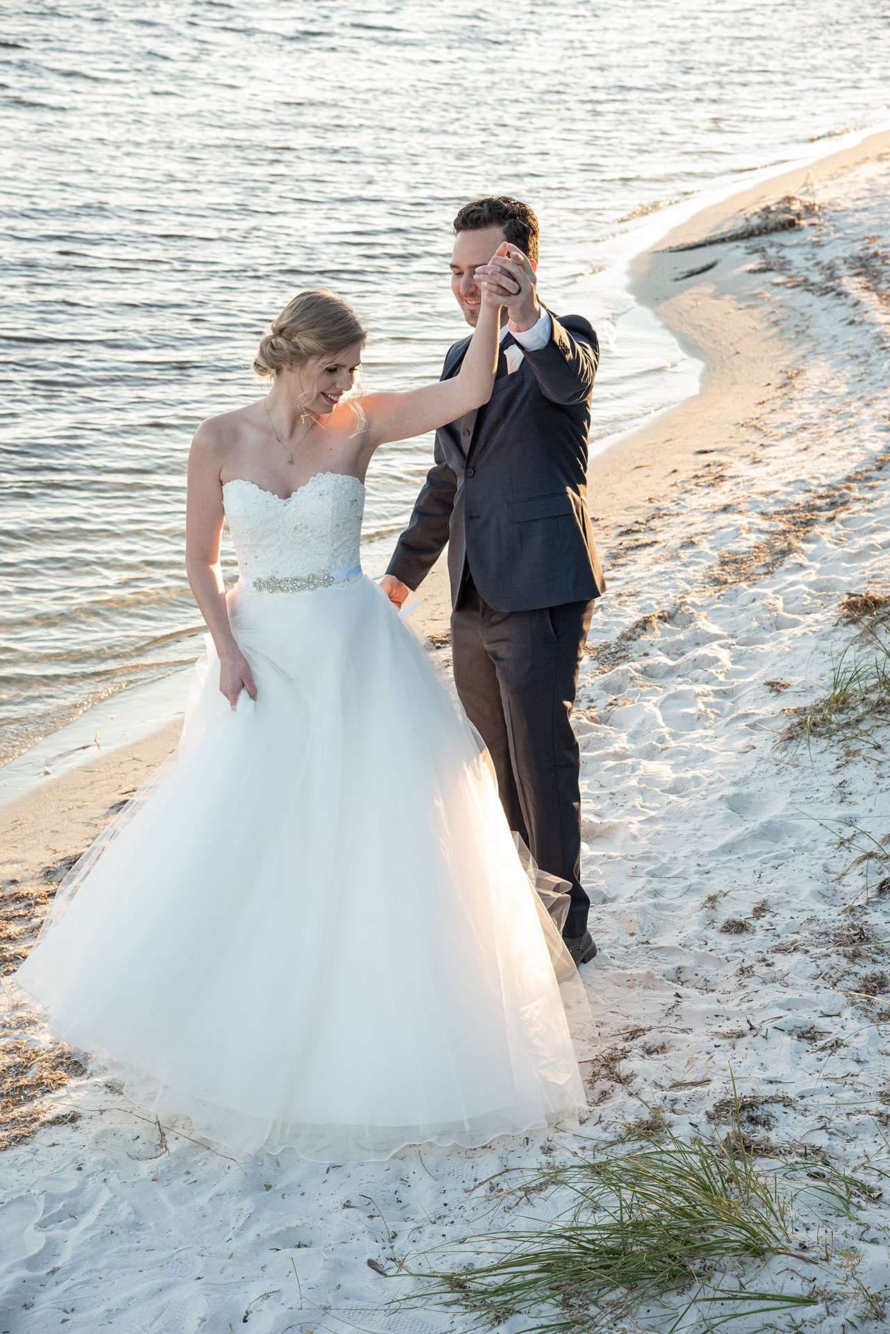 Bride in a white tulle gown twirling with her groom in a dark suit on a sandy shoreline