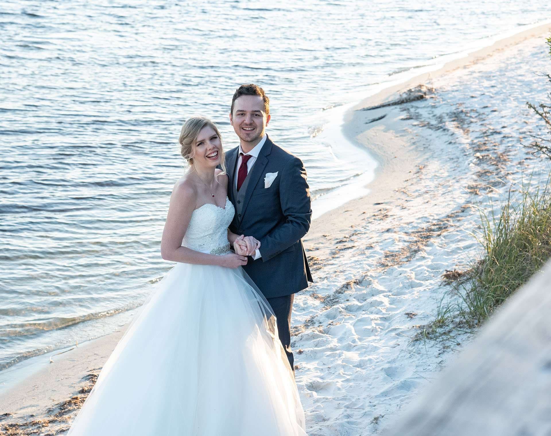 Smiling bride in a strapless white gown and groom in a gray suit holding hands on a sandy beach