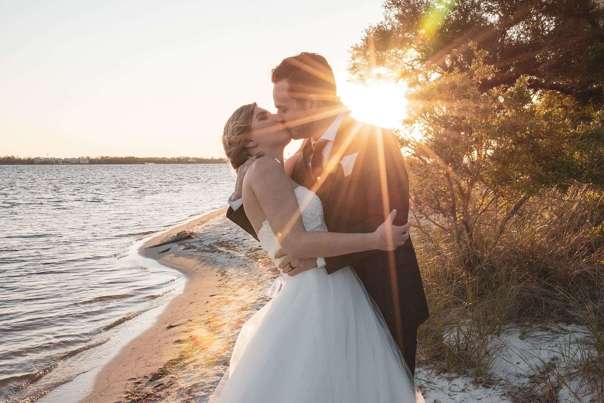 Couple kissing at sunset on a sandy shore, bride in a white gown, groom in a dark suit, sun flaring through trees