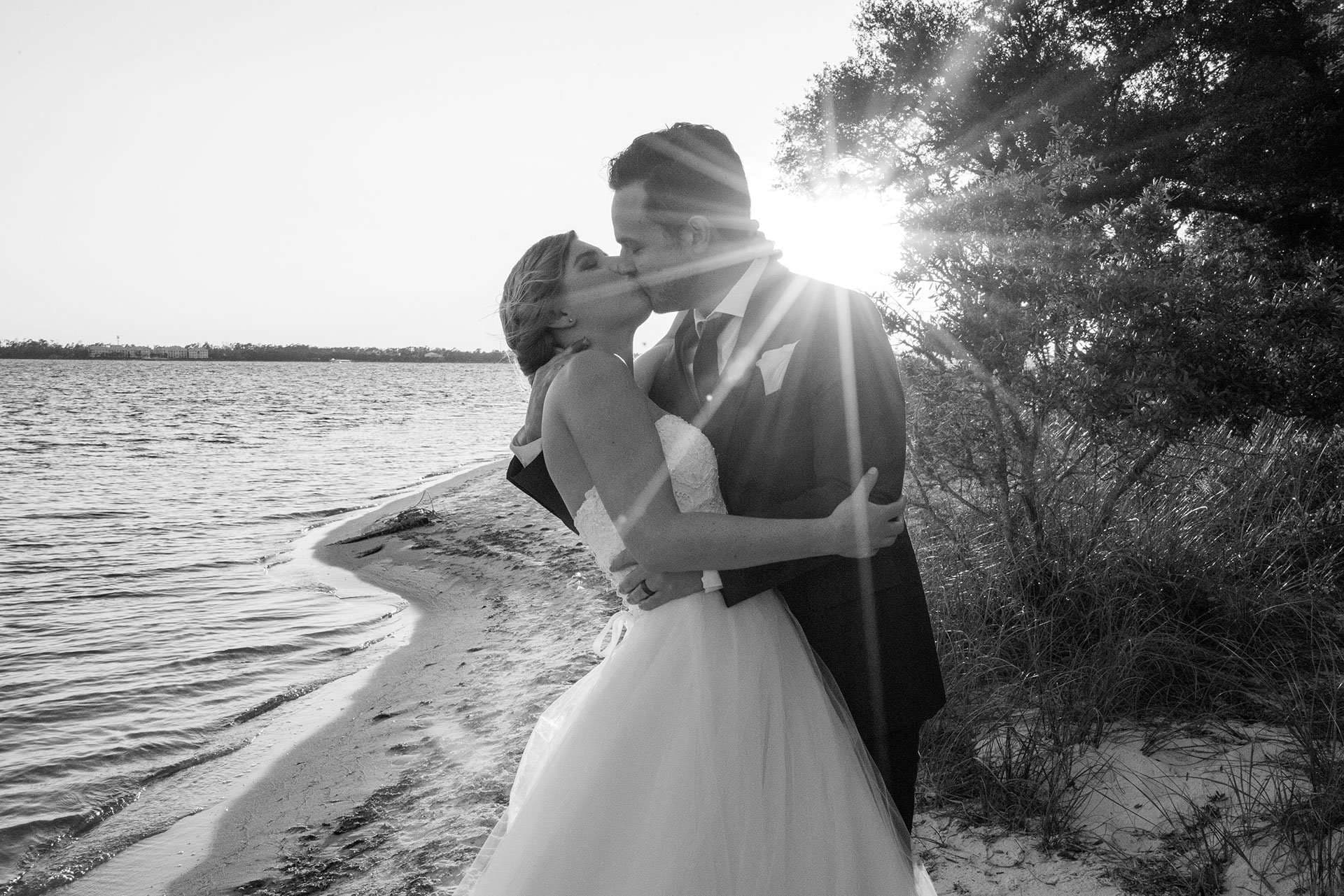 Black-and-white shot of a couple kissing on a sandy shoreline, bride in a tulle gown, sun flaring through trees