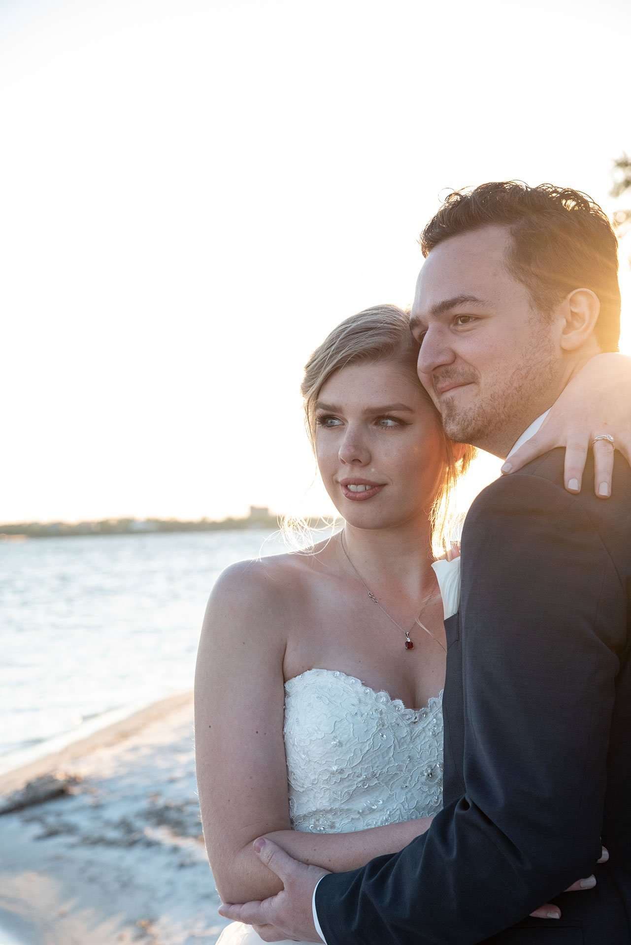 Bride in a strapless white lace gown leaning into her groom in a dark suit by the water at sunset