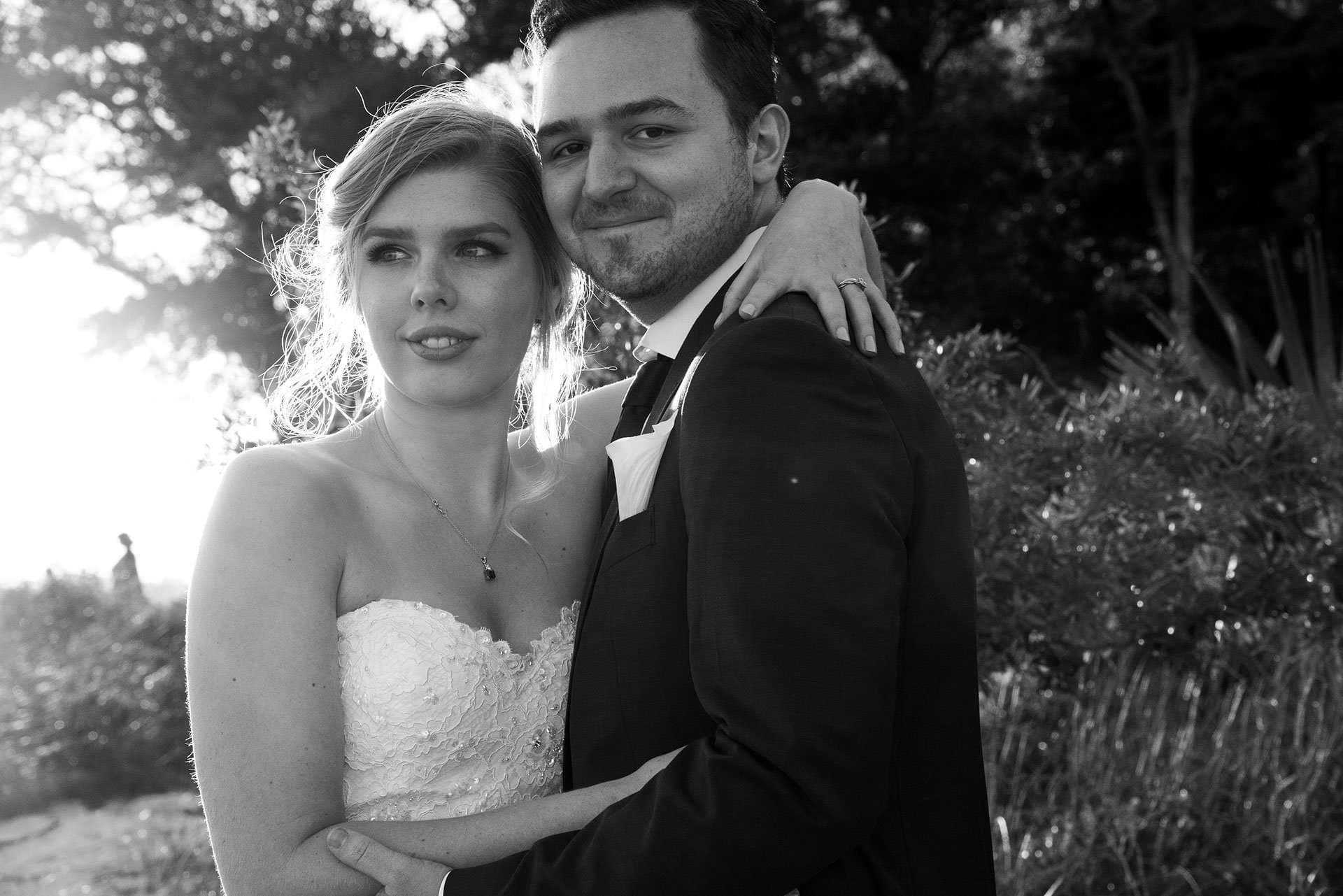 Black-and-white portrait of a bride in a strapless lace gown embraced by a smiling groom in a dark suit outdoors
