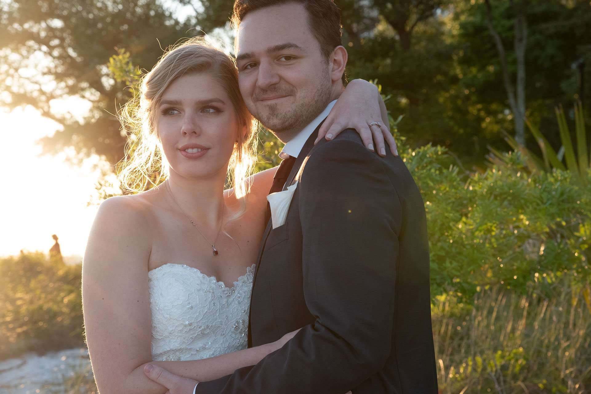 Bride in a strapless white lace gown embraced by a smiling groom in a dark suit, backlit by golden sunset light