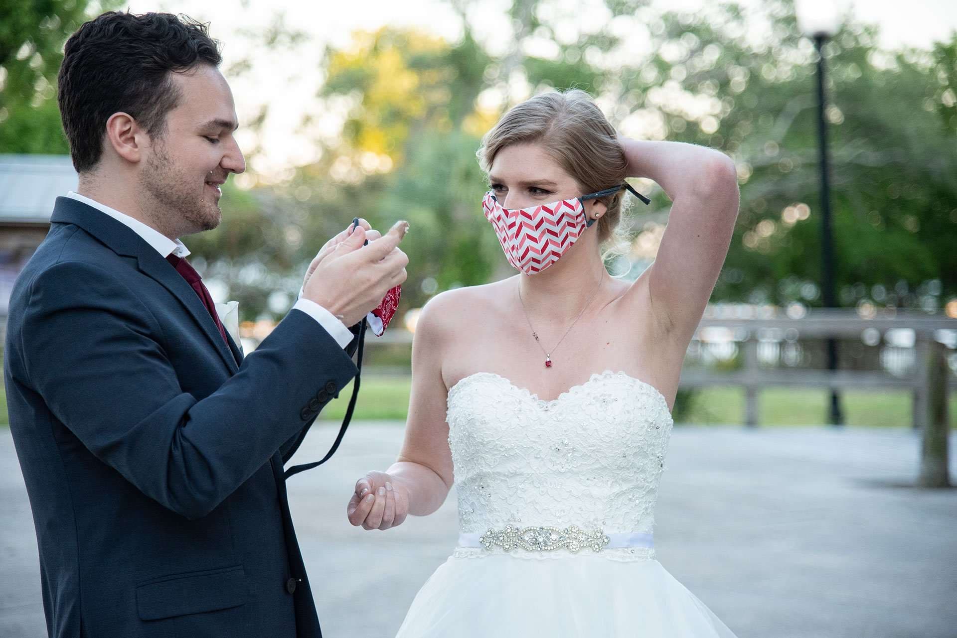 Bride in a strapless white gown adjusts her red chevron face mask while the groom in a navy suit smiles beside her