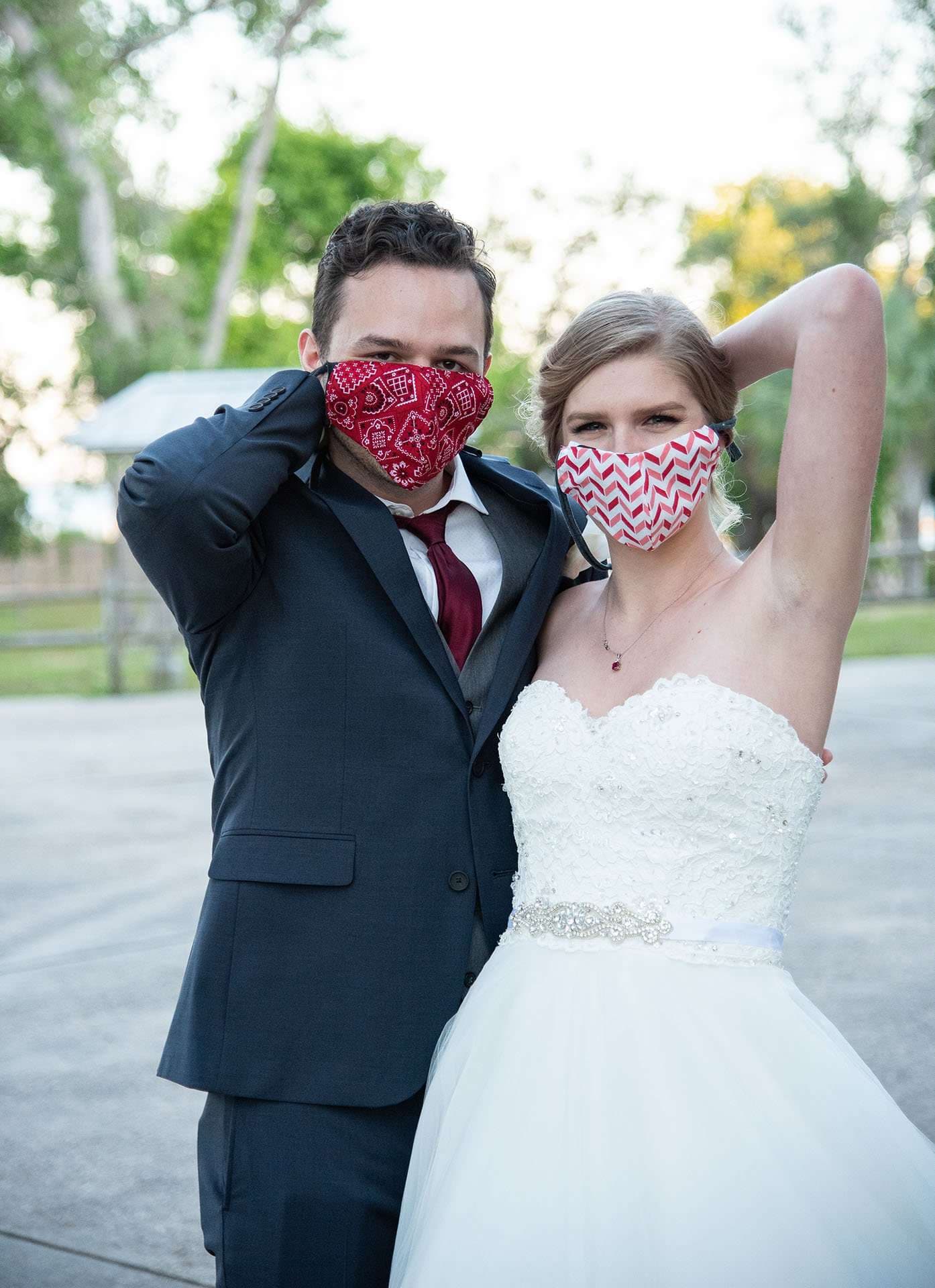 Bride and groom in red patterned face masks posing close together outdoors, each with one arm raised behind the head