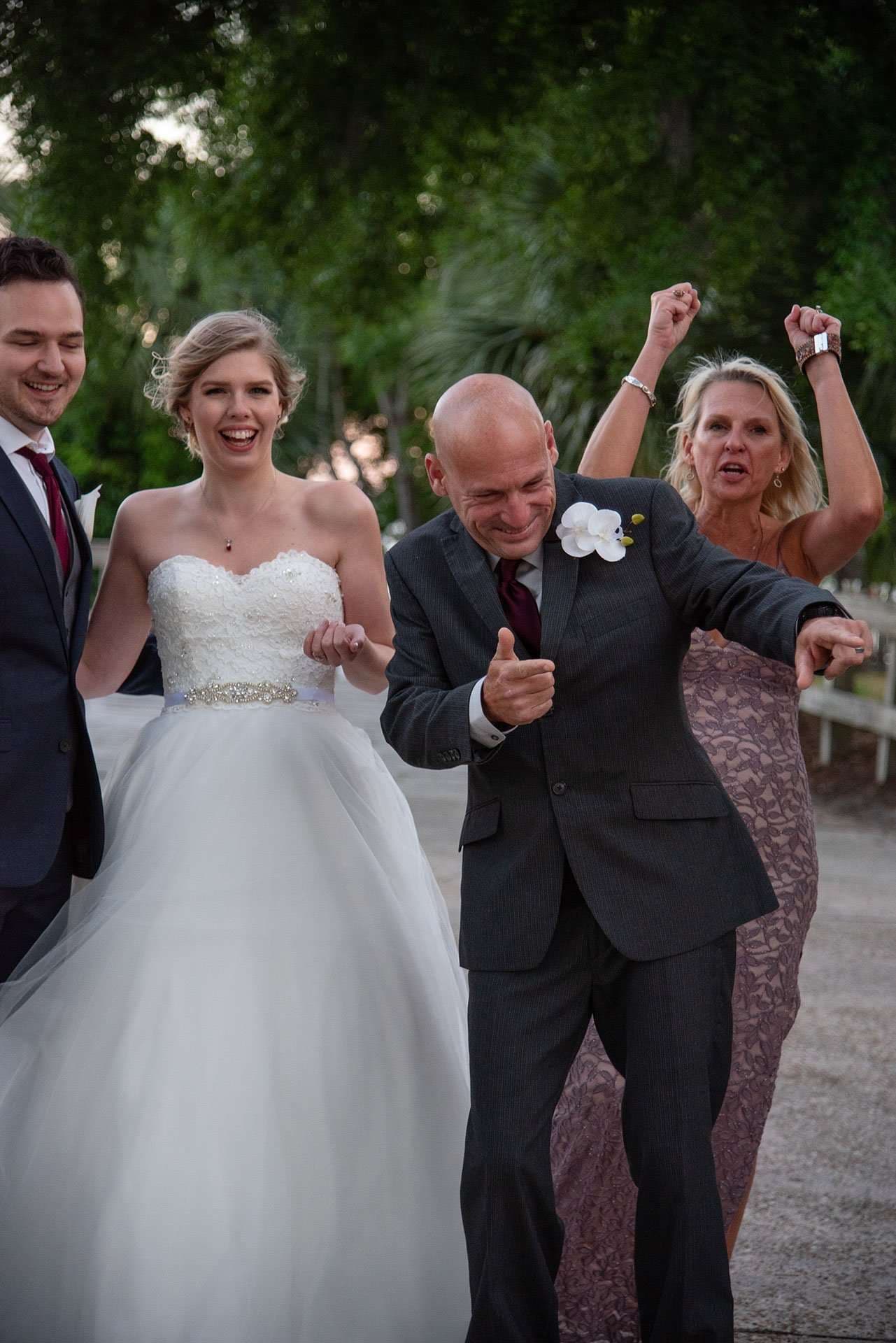 Bride in a white strapless gown laughs as a groom and a woman in a lace dress dance outdoors