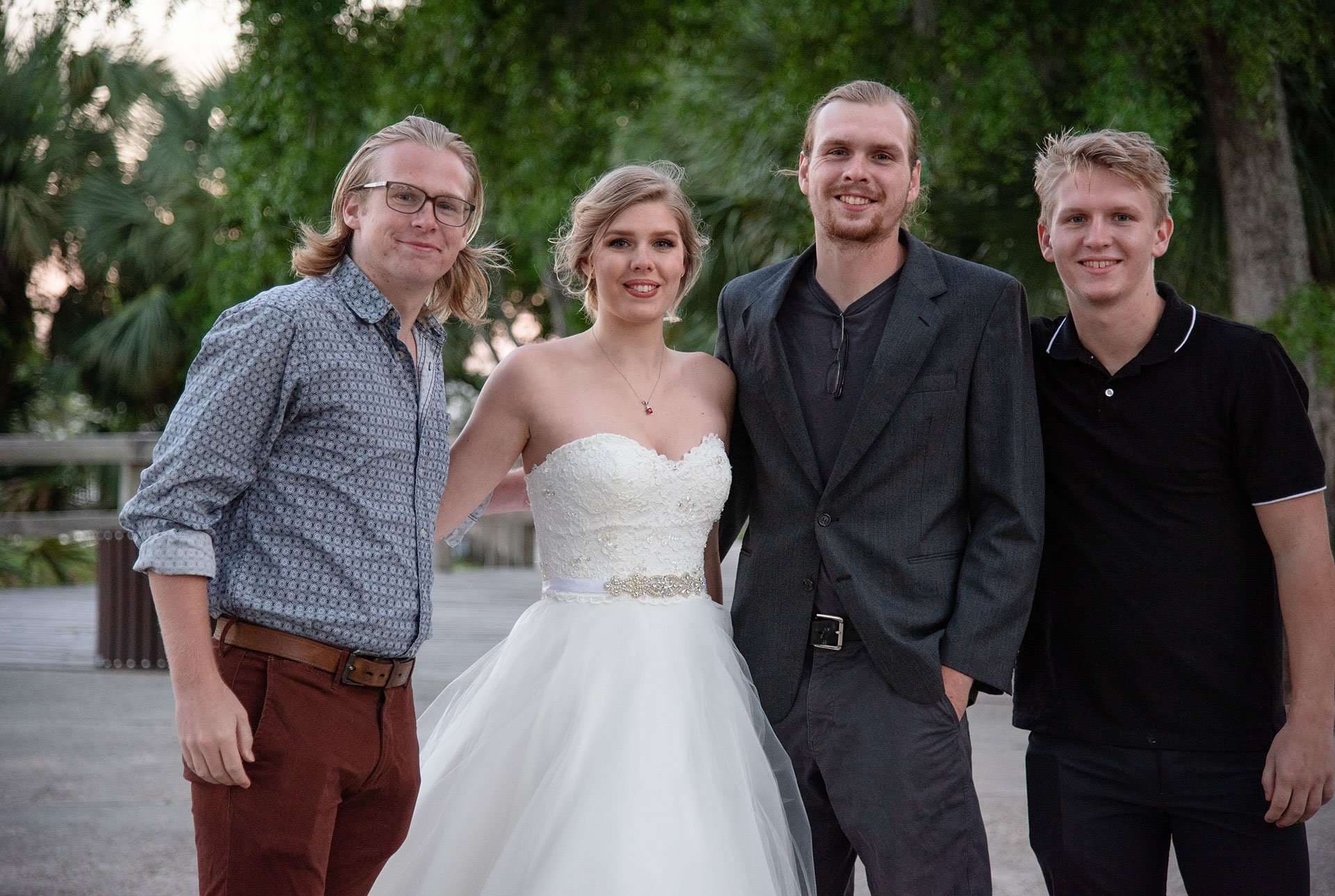 Bride in a strapless white gown posing outdoors with three smiling men in casual clothes among green trees